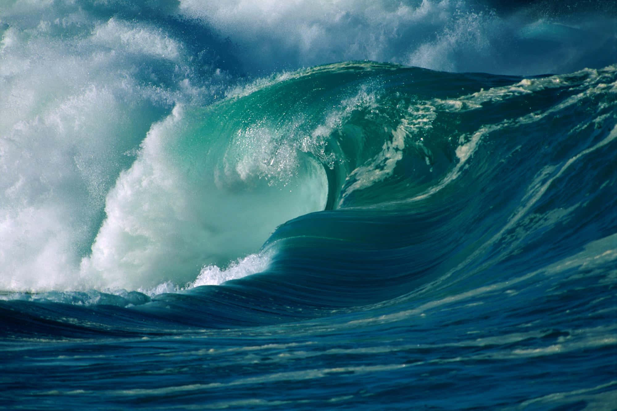 waves 桌布 壁紙 — 巨大的藍綠色海浪捲成一個完美的筒狀，伴隨著泡沫般的白色浪花，映襯著暴風雨的天空。