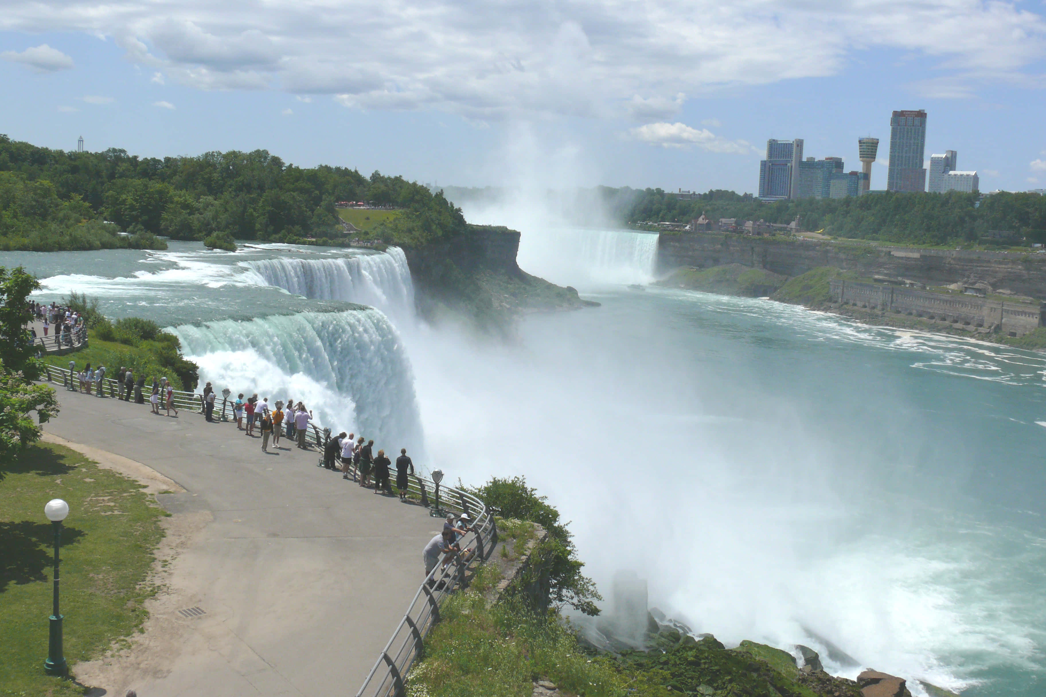 Niagara Falls 桌布 壁紙 — 巨大的藍綠色瀑布伴隨著水霧咆哮，遊客們在部分多雲的天空下沿著觀景路排隊，遠處是城市摩天大樓。