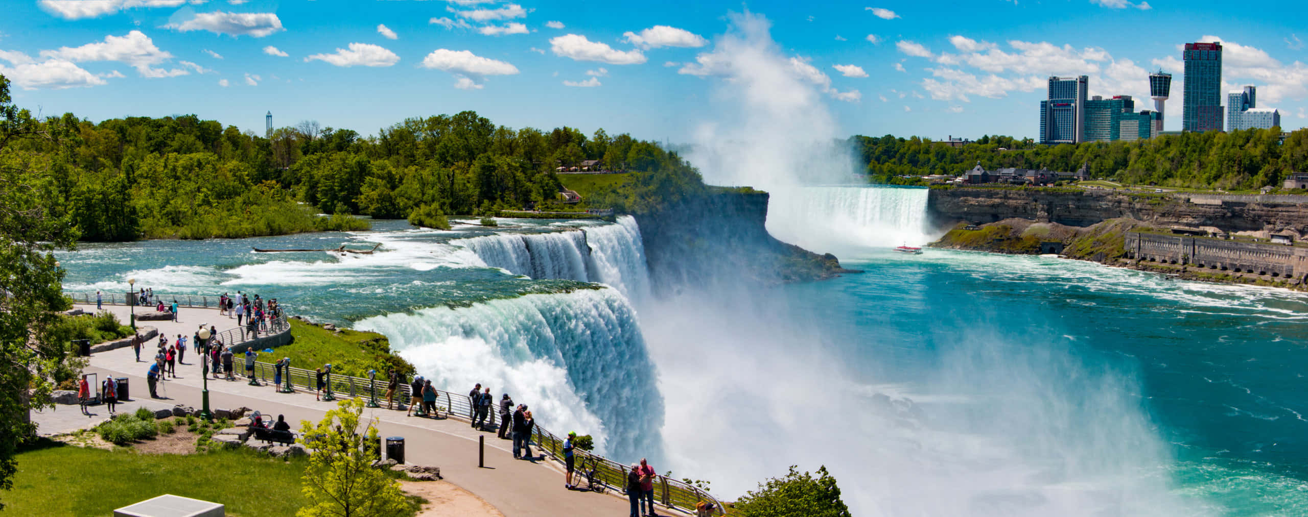 Niagara Falls Canada 桌布 壁紙 — 巨大的藍綠色瀑布在明亮的藍天和散落的雲朵下咆哮，遊客沿著觀景步道行走，遠處是城市天際線。