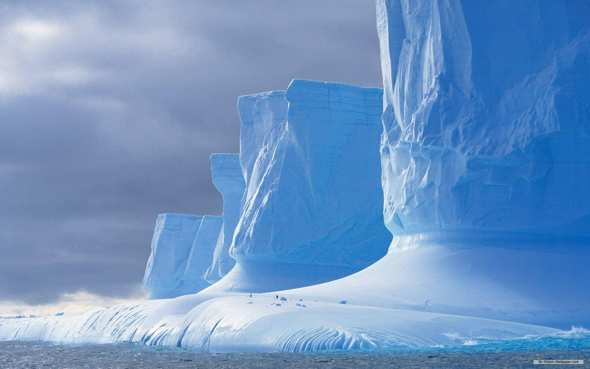 Antarctica 桌布 壁紙 — 巨大的藍色冰山從冰冷的海水中升起，在戲劇性的多雲天空下，有微小的人物作為比例參考。