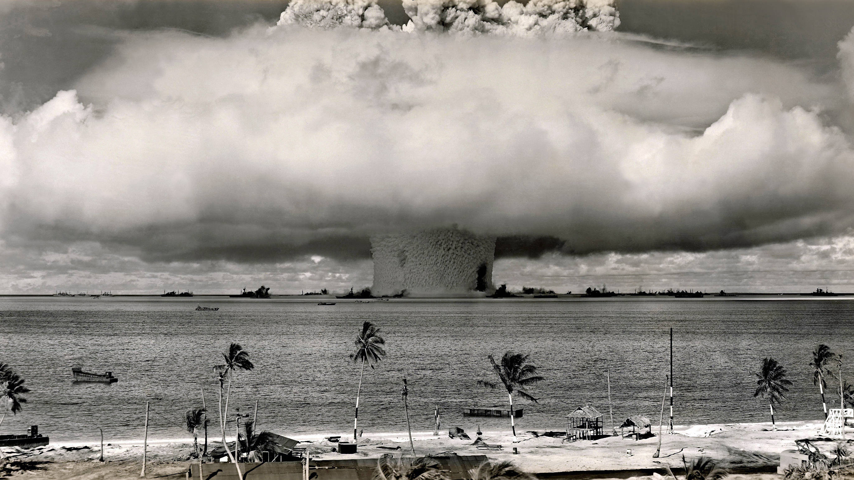 nuclear explosion 桌布 壁紙 — 巨大的蘑菇雲在熱帶海灘上升起，遠處有棕櫚樹和船隻。