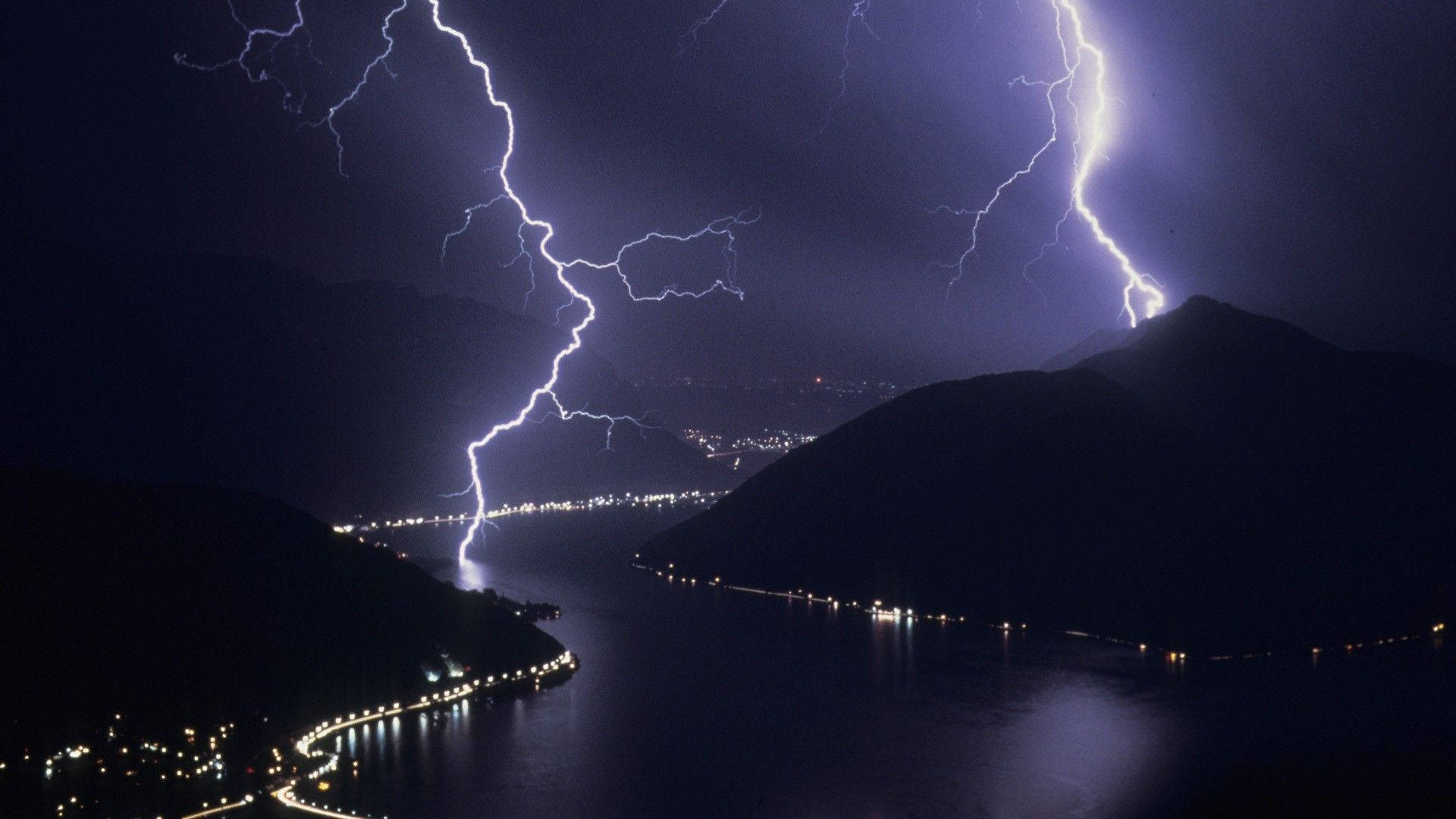 Aesthetic Lightning 桌布 壁紙 — 巨大的閃電照亮了夜晚黑暗的山湖，城市燈光沿著海岸線閃爍。