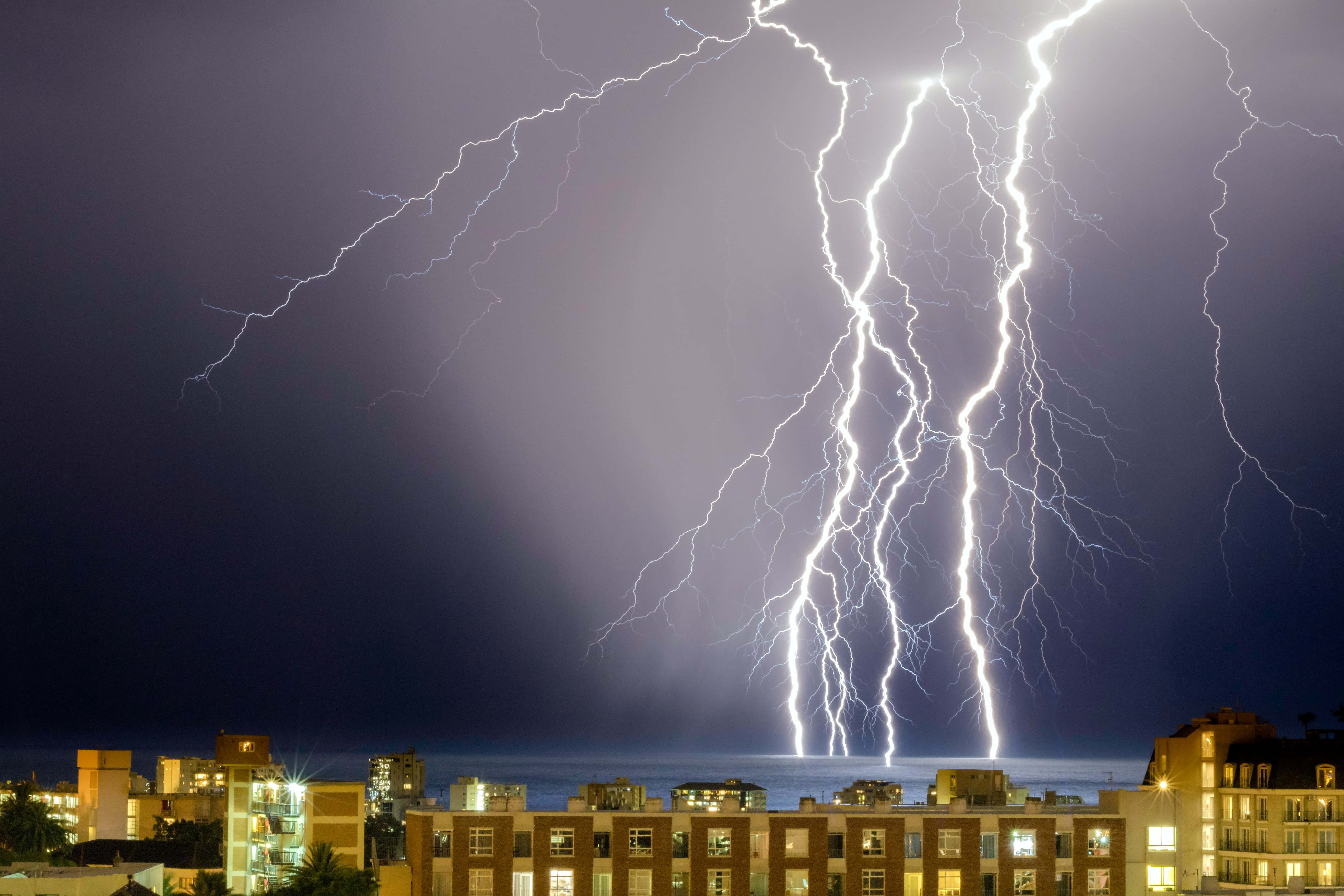 電力 桌布 壁紙 — 巨大的閃電照亮了暴風雨夜晚的天空，下方是發光的城市建築。