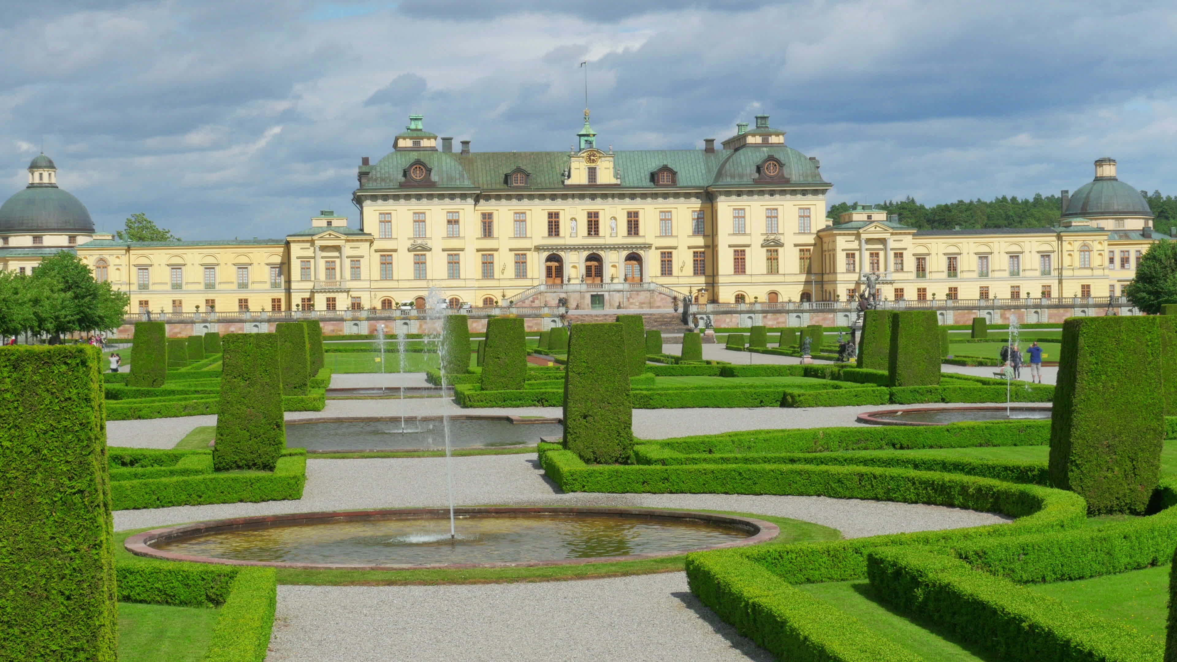 Drottningholm Palace 桌布 壁紙 — 巨大的黃色宮殿，綠色圓頂，俯瞰著對稱的花園，園內有修剪整齊的樹籬、噴泉和礫石小徑，天空多雲。