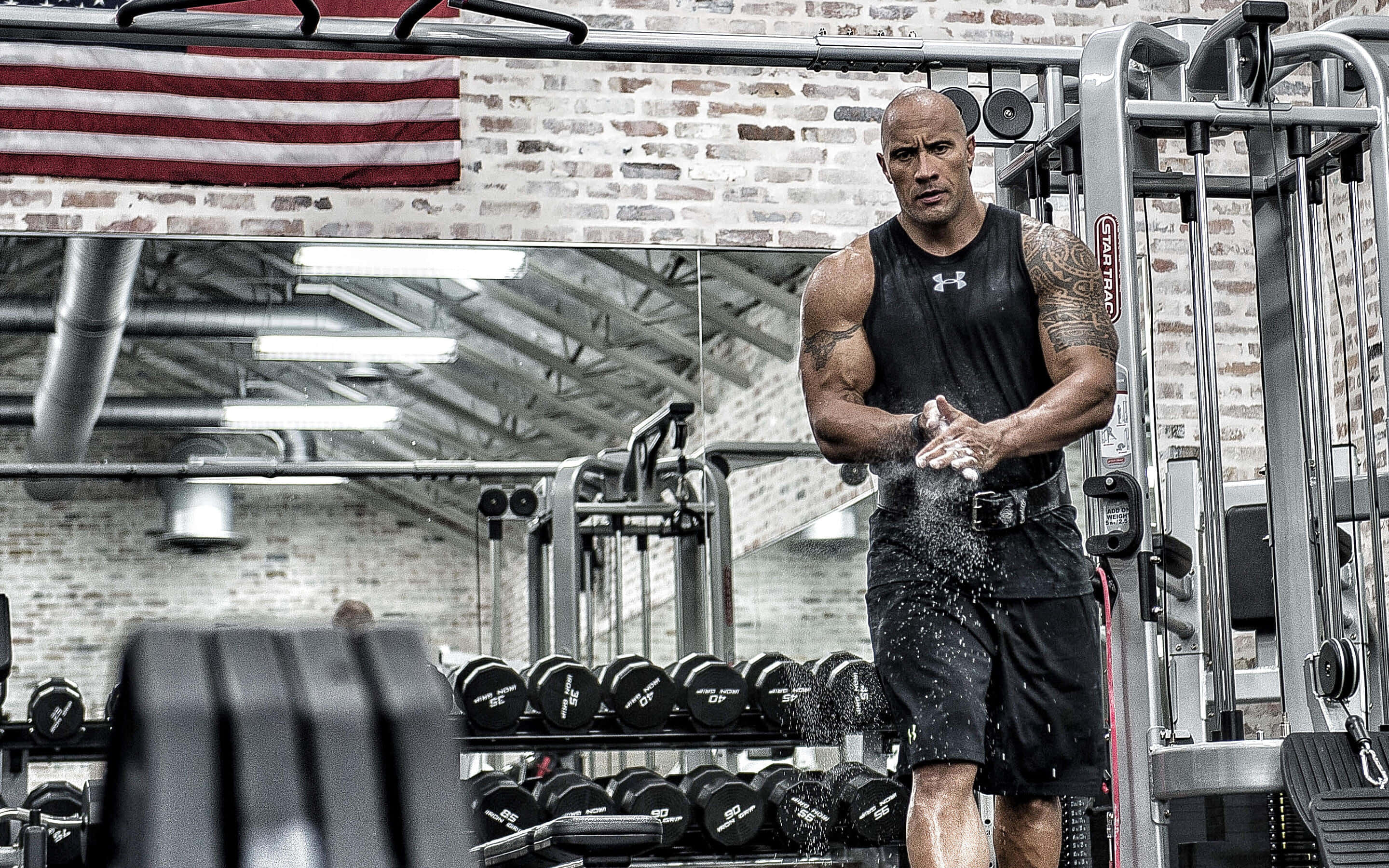 The Rock 桌布 壁紙 — 巨石強森在一個粗糙的健身房中，將粉筆灰撒向空中，周圍是啞鈴和重量訓練機。