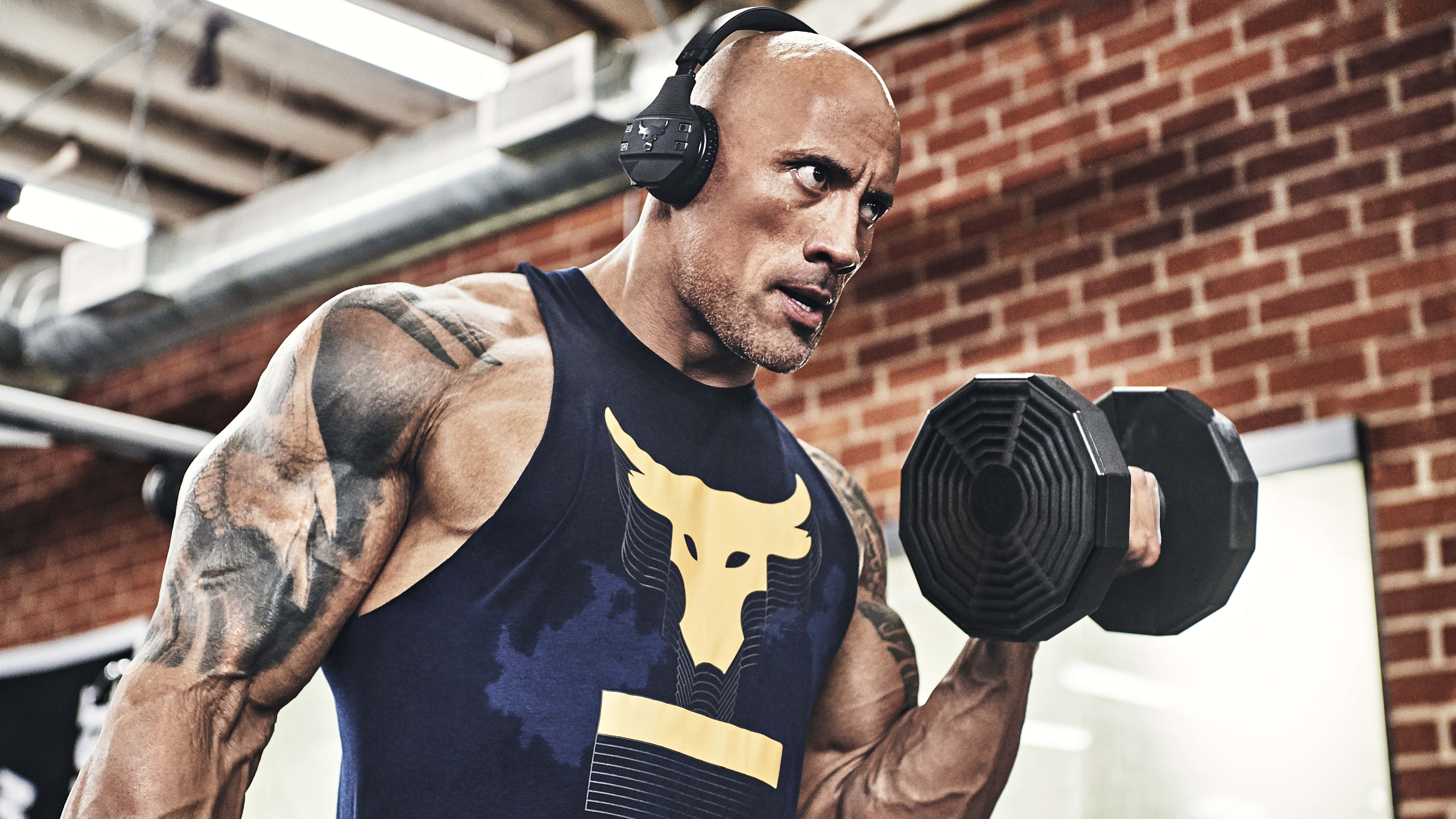 The Rock 桌布 壁紙 — 巨石強森在健身房中用力舉起啞鈴，牆壁是裸露的磚牆，搭配工業風照明。
