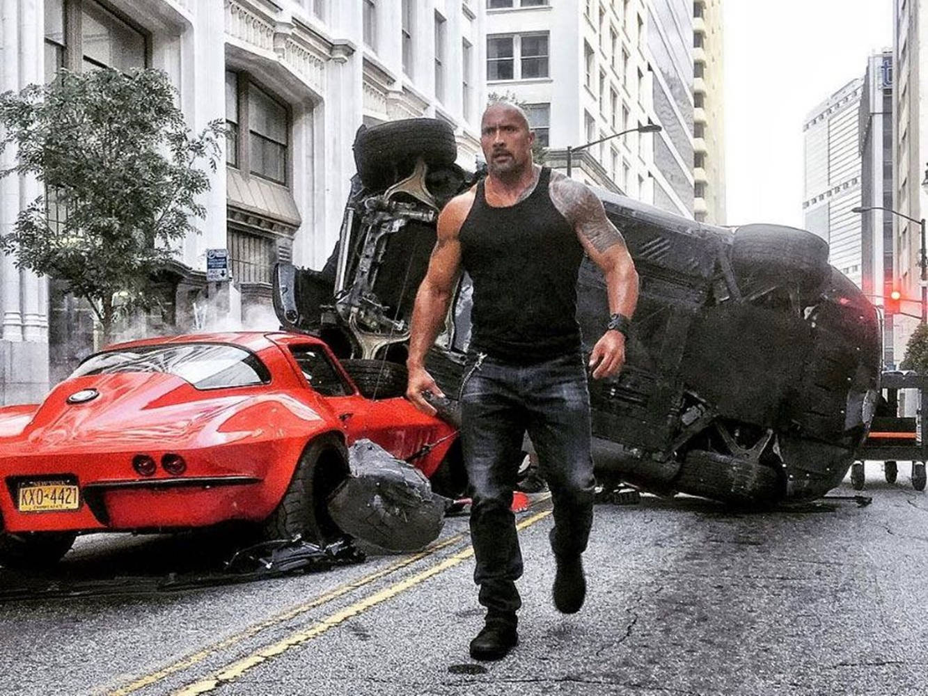 The Rock 桌布 壁紙 — 巨石強森在城市混亂中，走過一輛紅色跑車和一輛翻覆的黑色車輛，在城市街道上大步前行。