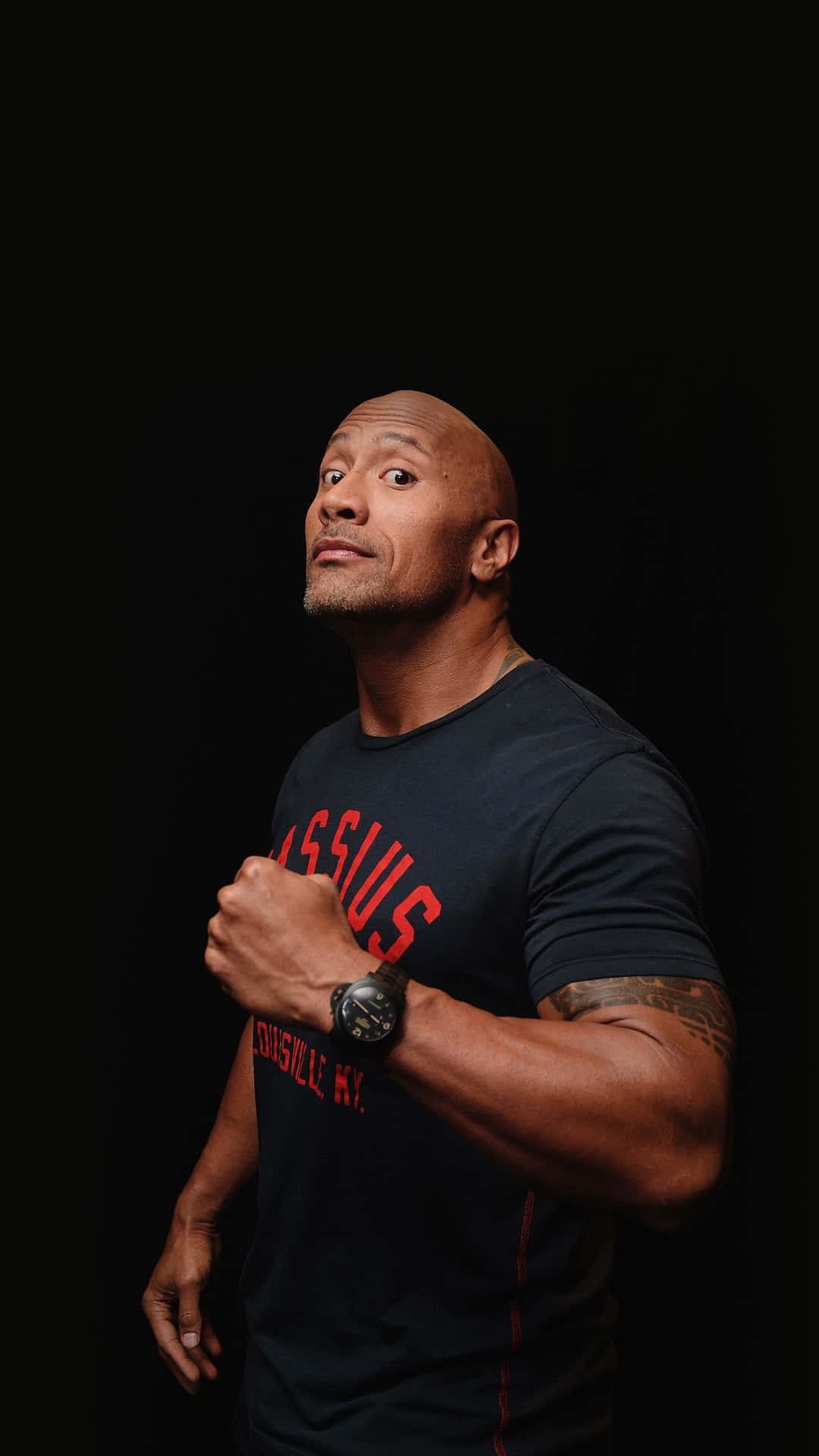 The Rock 桌布 壁紙 — 巨石強森在純黑背景前睜大雙眼向上看，身穿印有紅色文字的深色T恤。