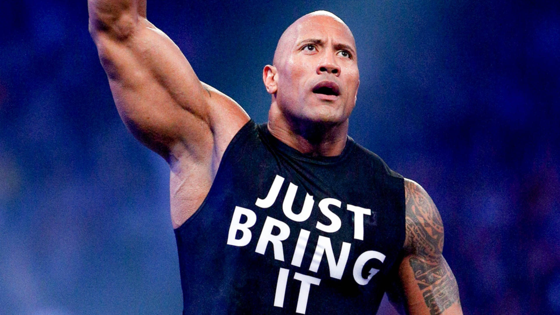The Rock 桌布 壁紙 — 巨石強森在藍色模糊背景前，穿著黑色背心，上面有白色字寫著 just bring it，展示他肌肉發達的手臂。