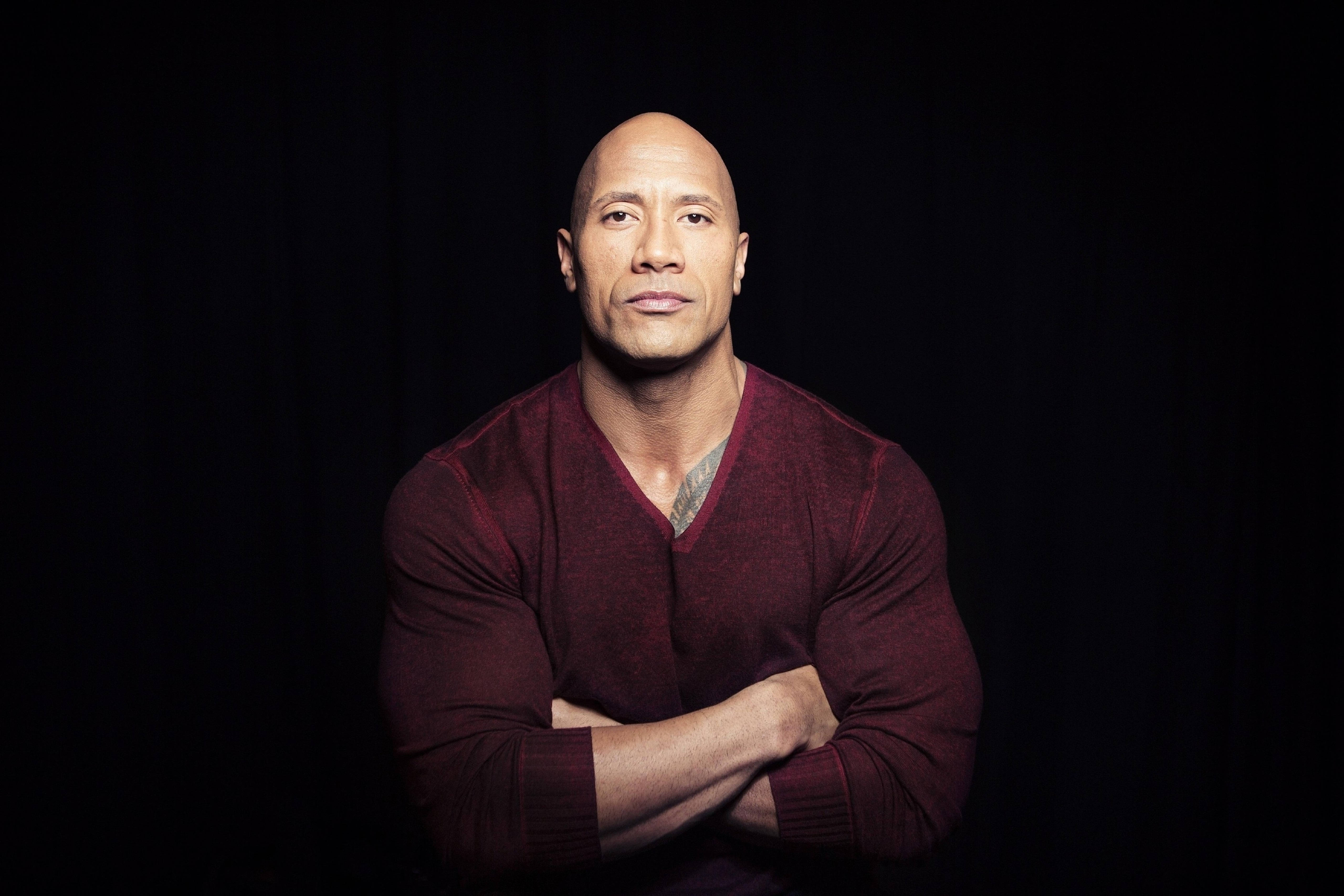 Dwayne 桌布 壁紙 — 巨石強森自信地交叉雙臂，站在純黑色背景前，身穿栗色毛衣。