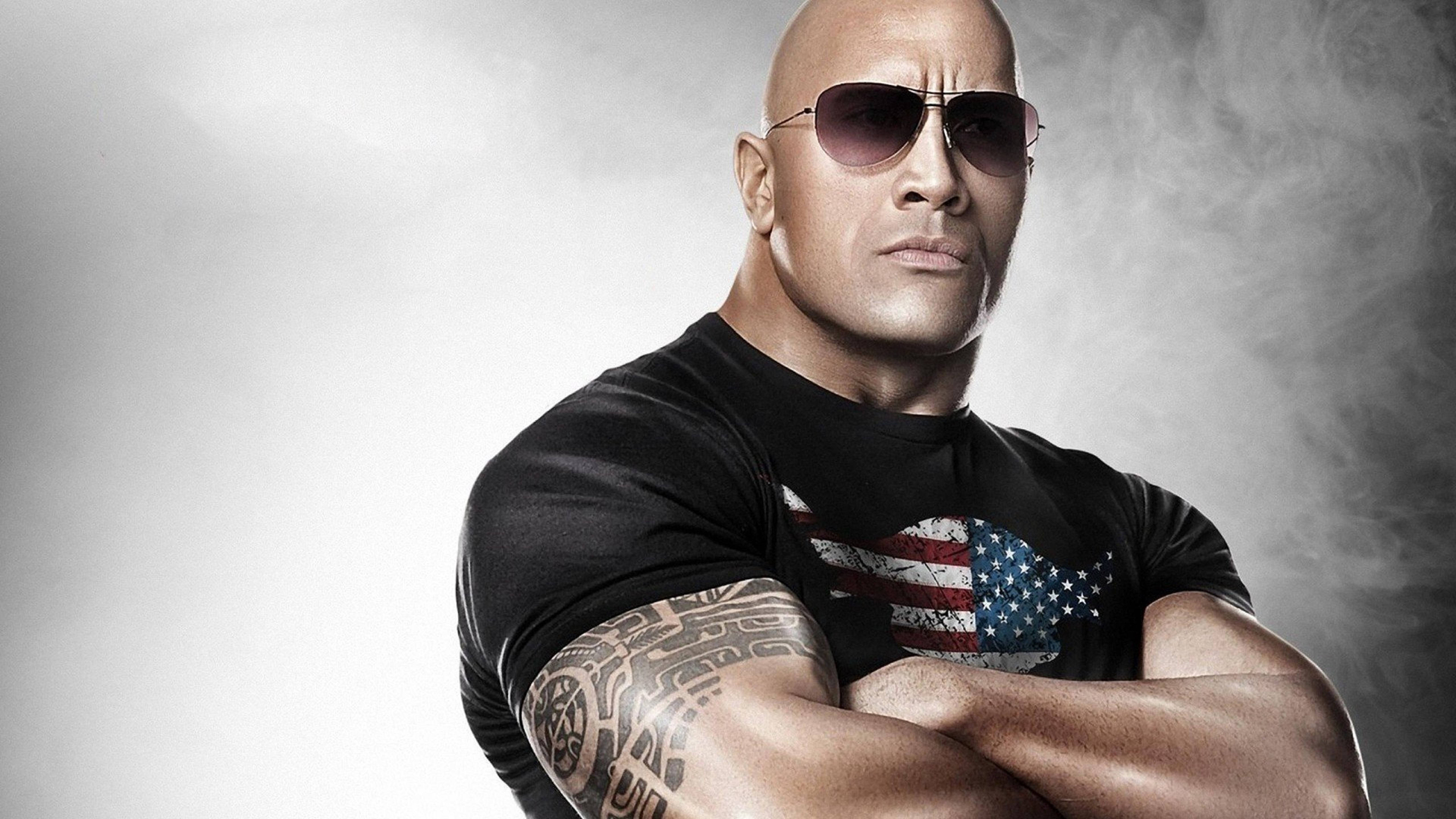 Dwayne 桌布 壁紙 — 巨石強森自信地交叉雙臂站立，戴著太陽眼鏡，身穿帶有美國國旗圖案的黑色T恤，背景是煙霧灰色。