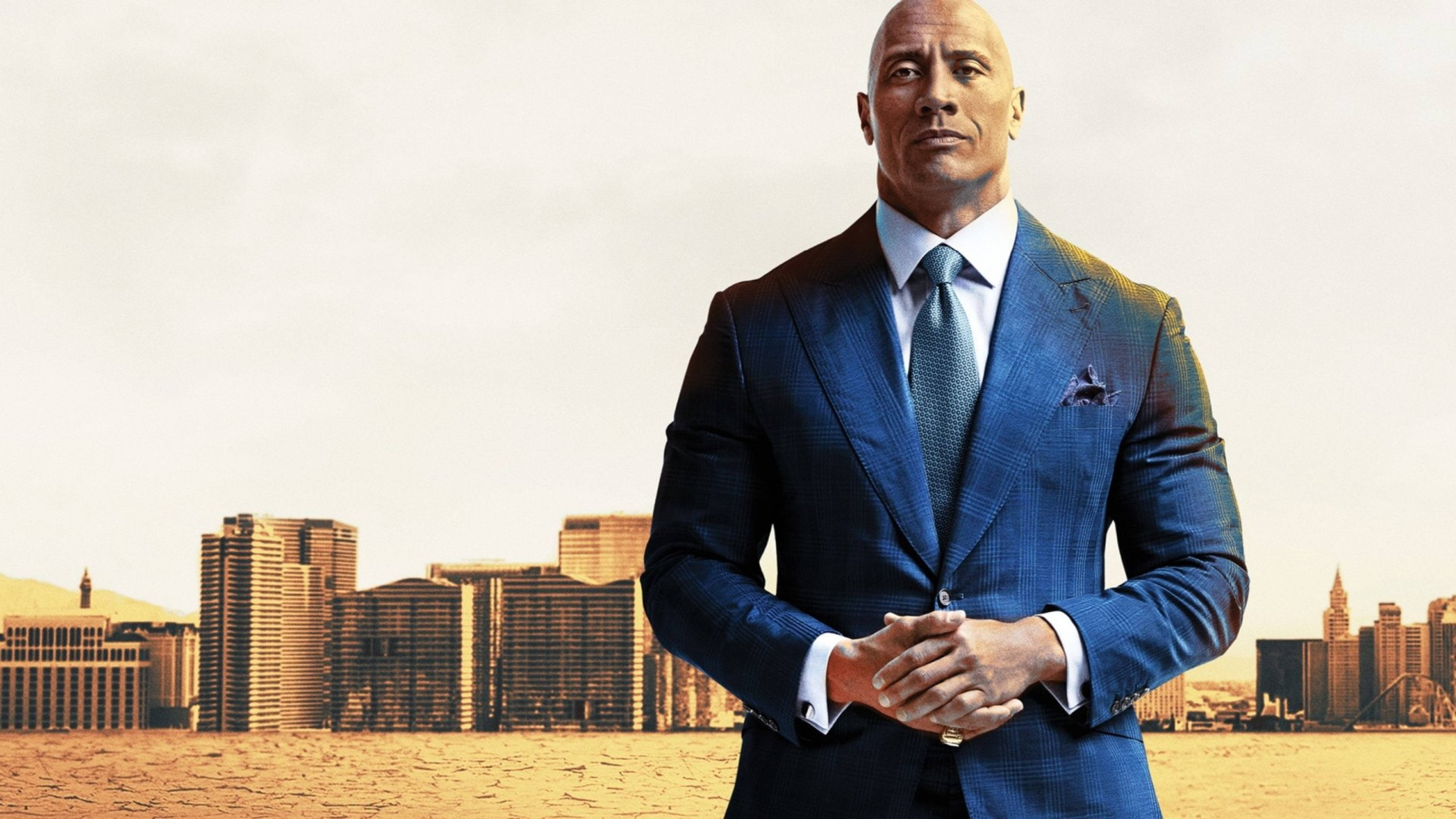 Dwayne 桌布 壁紙 — 巨石強森自信地穿著一套俐落的藍色西裝，背景是模糊的城市天際線和現代建築。