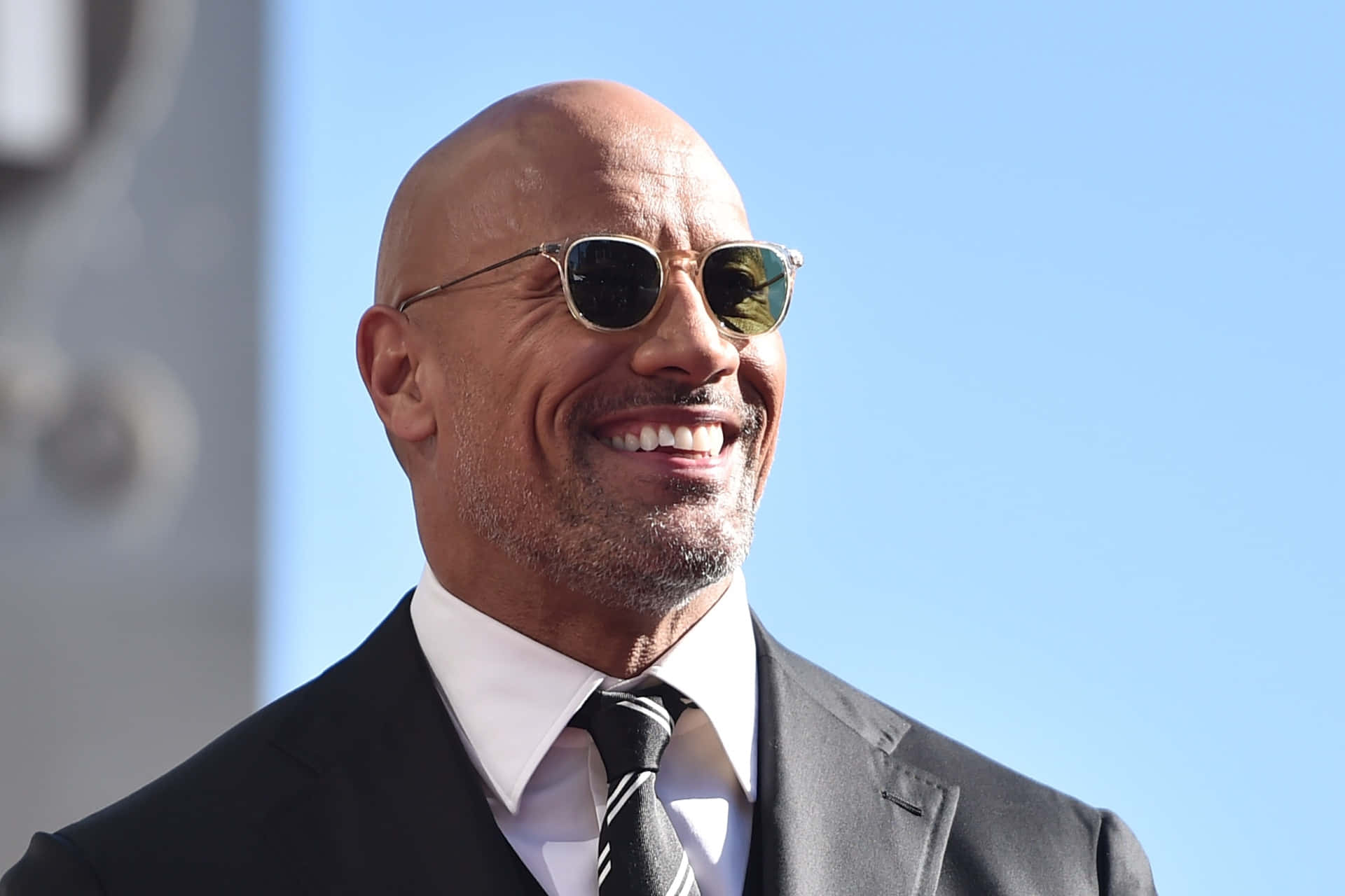 Celebrity 桌布 壁紙 — 巨石強森身穿俐落西裝和太陽眼鏡，在晴朗的藍天下閃耀。