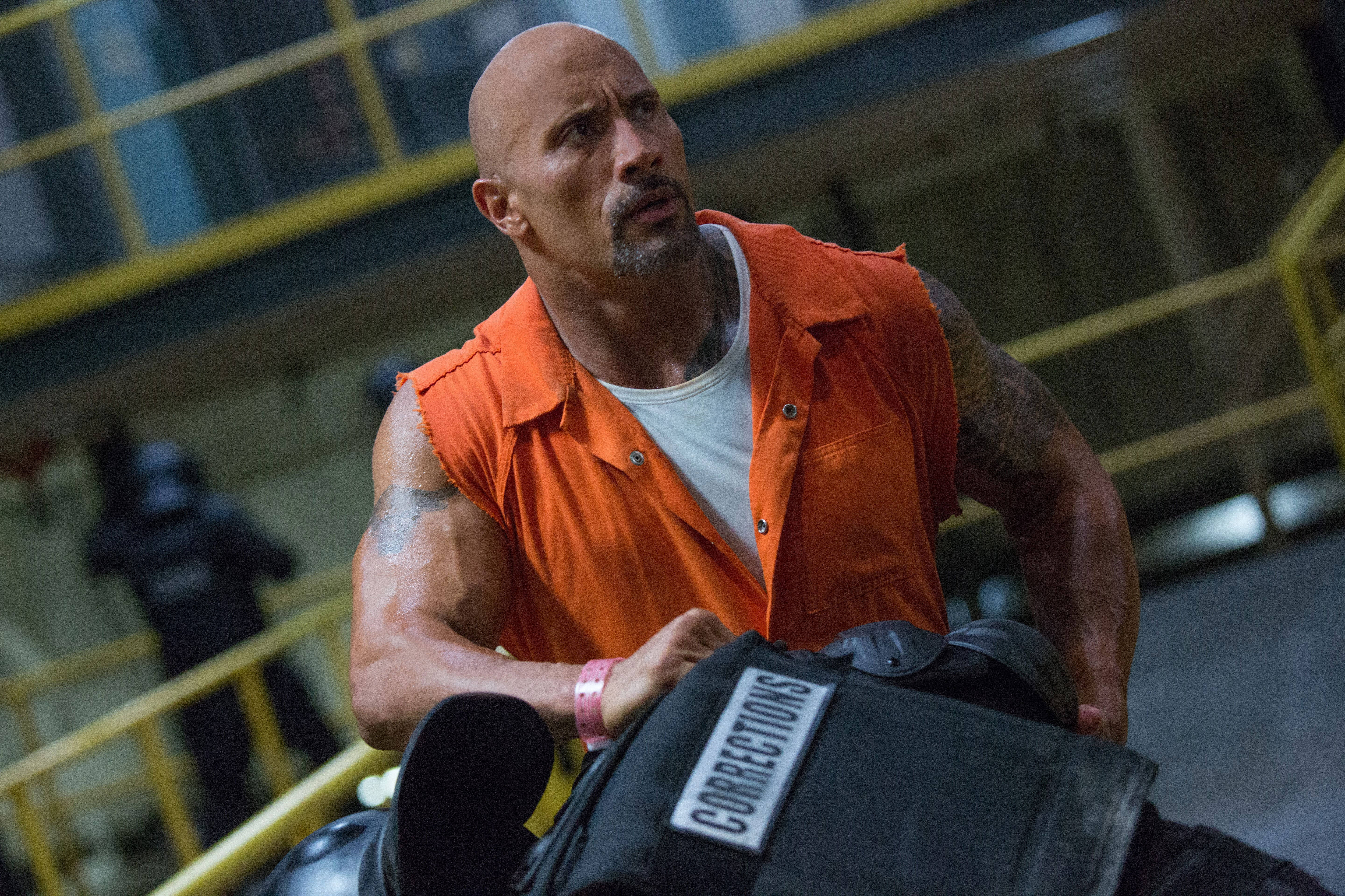 The Rock 桌布 壁紙 — 巨石強森身穿橙色監獄連身衣，在陰暗的工業設施中用力推動沉重的黑色矯正推車。
