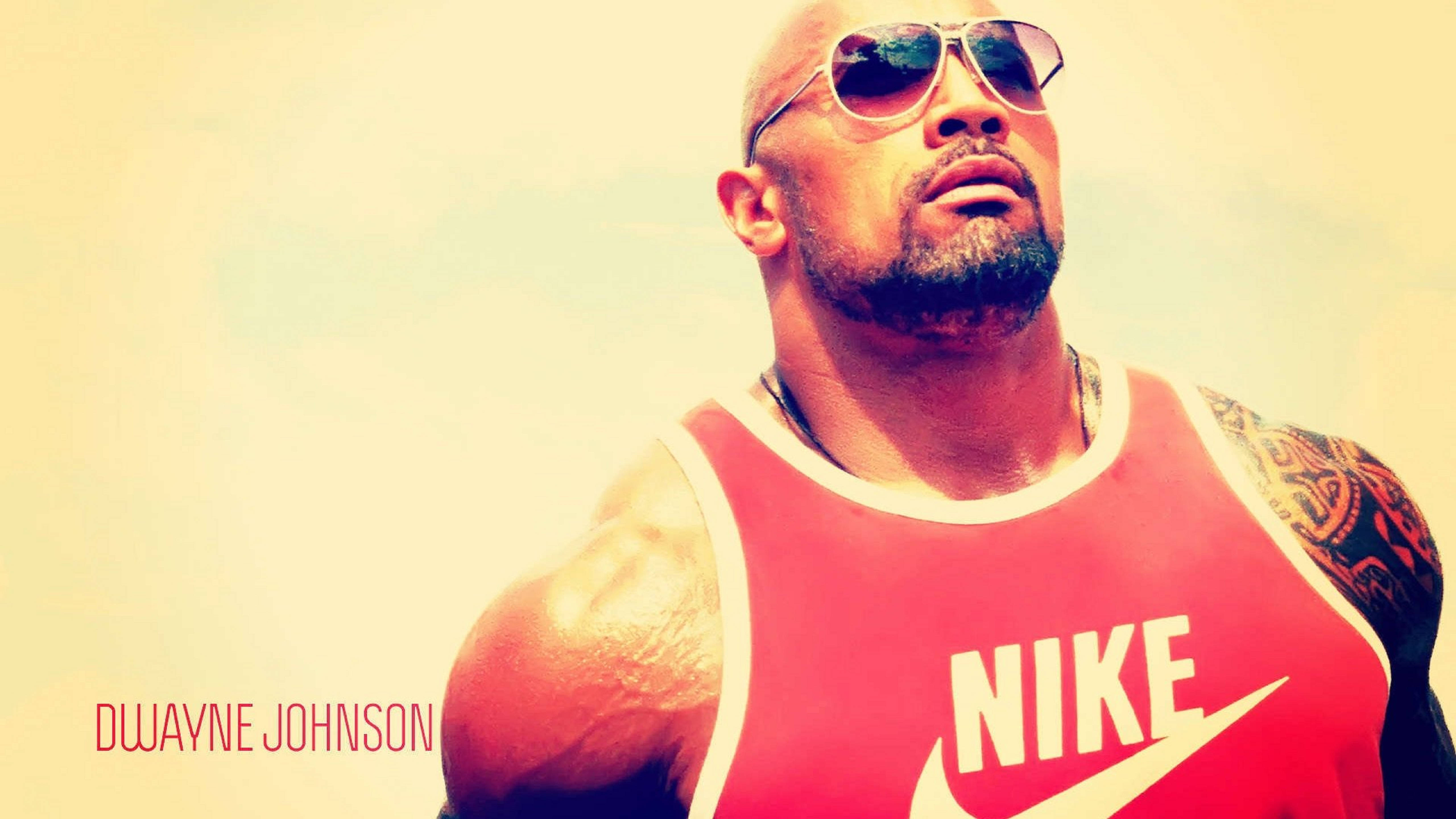 Dwayne 桌布 壁紙 — 巨石強森身穿紅色Nike背心及飛行員太陽眼鏡，在溫暖朦朧的背景前自信地凝視。