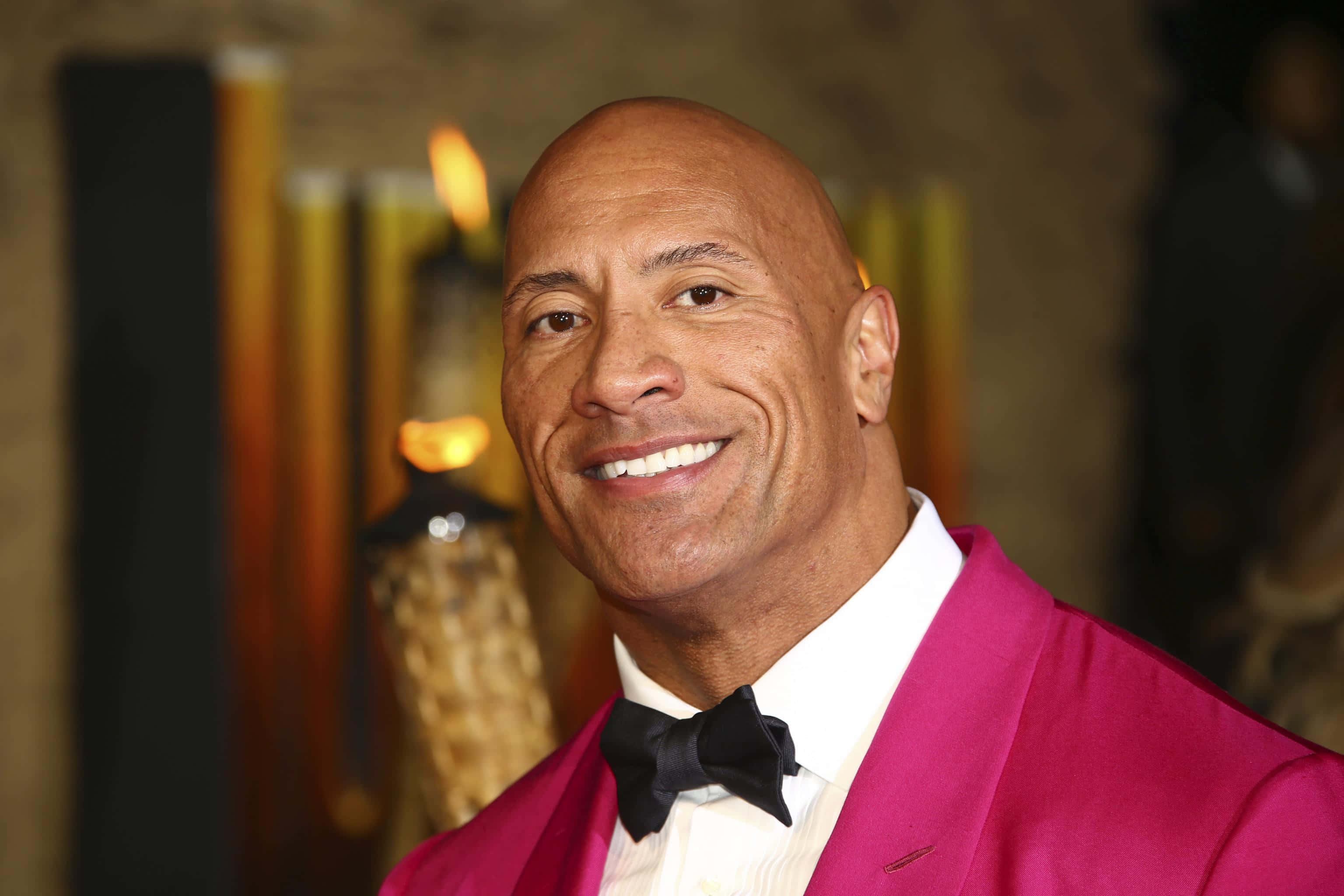 Celebrity 桌布 壁紙 — 巨石強森身穿鮮豔的洋紅色西裝外套搭配黑色蝴蝶結，在溫暖的燭光背景下閃耀。