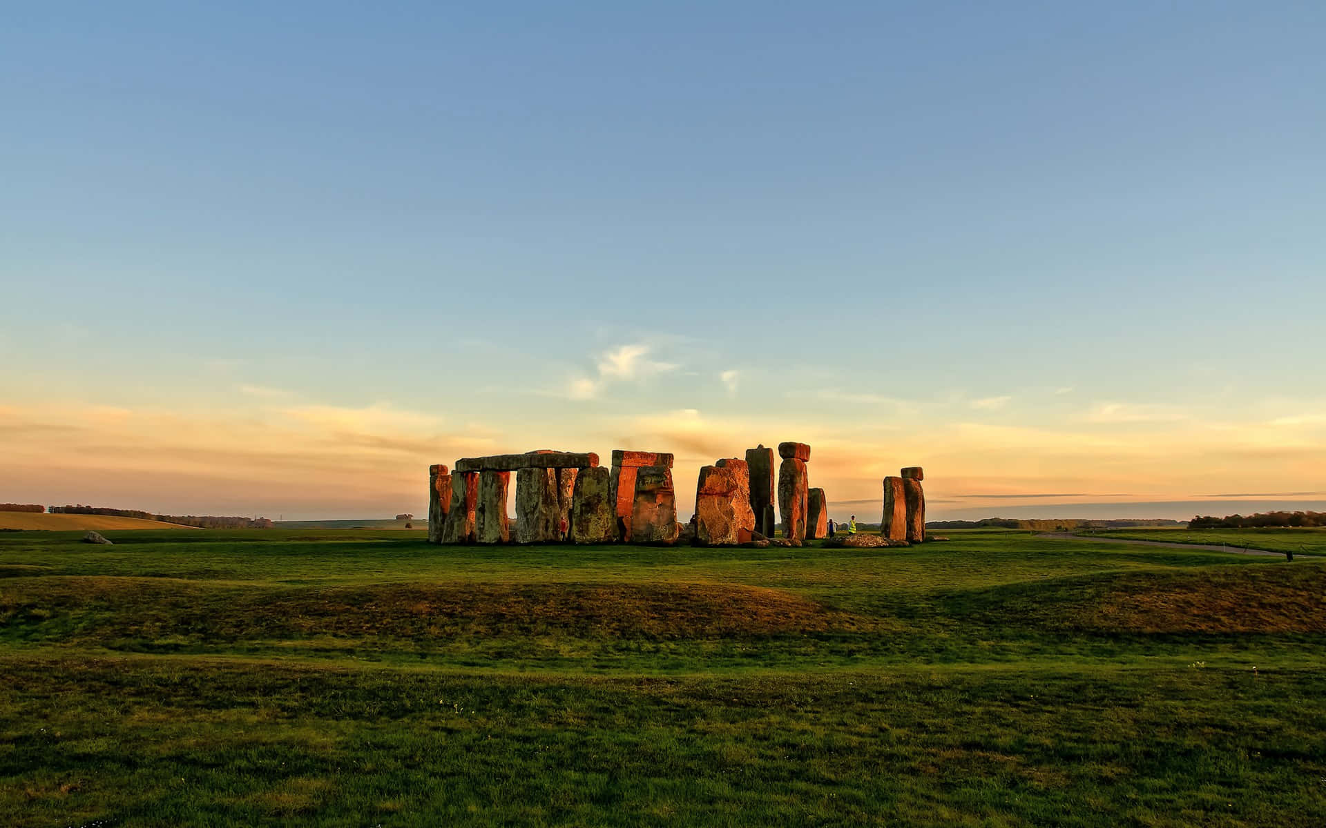 England 桌布 壁紙 — 巨石陣的古老立石在金色夕陽下閃耀，映襯著連綿起伏的綠色山丘上寧靜的藍天。