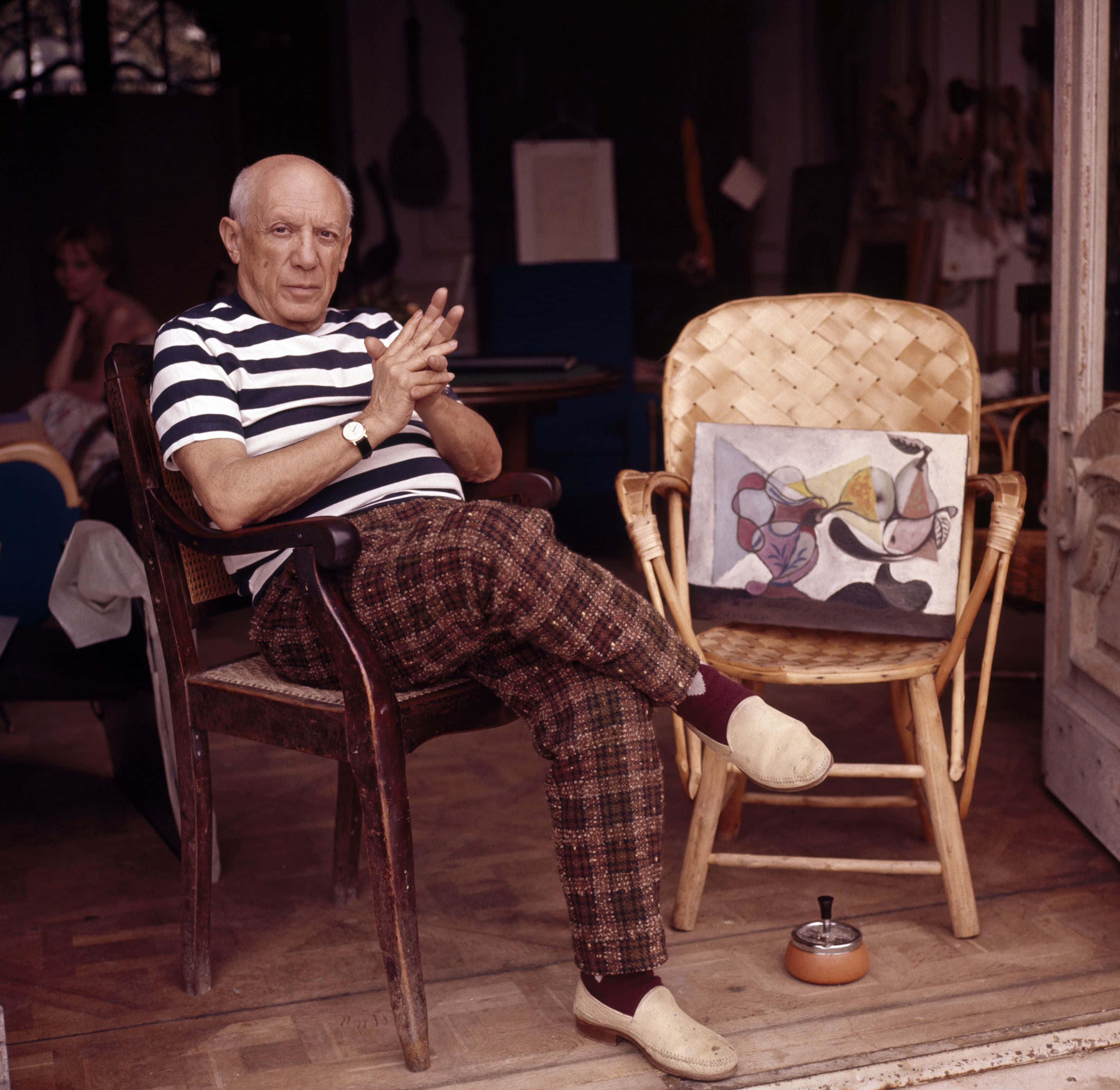 Picasso 桌布 壁紙 — 巴勃羅·畢卡索放鬆地坐在他凌亂工作室的木椅上，附近一張編織椅上放著他的抽象藝術品。