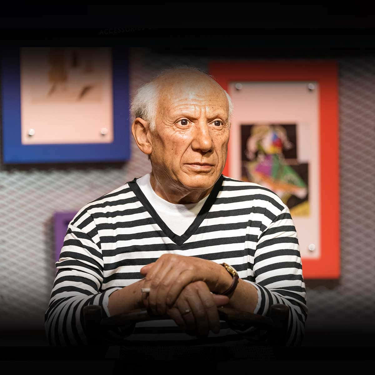 Picasso 桌布 壁紙 — 巴勃羅·畢卡索的逼真蠟像身穿其標誌性條紋襯衫，雙手交握而坐，周圍是質感牆壁上色彩繽紛的藝術作品。