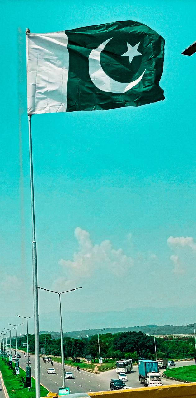 巴基斯坦國旗 桌布 壁紙 — 巴基斯坦國旗在高高的旗桿頂端自豪地飄揚，背景是充滿活力的藍綠色天空，下方是繁忙的高速公路和遠處的山丘。