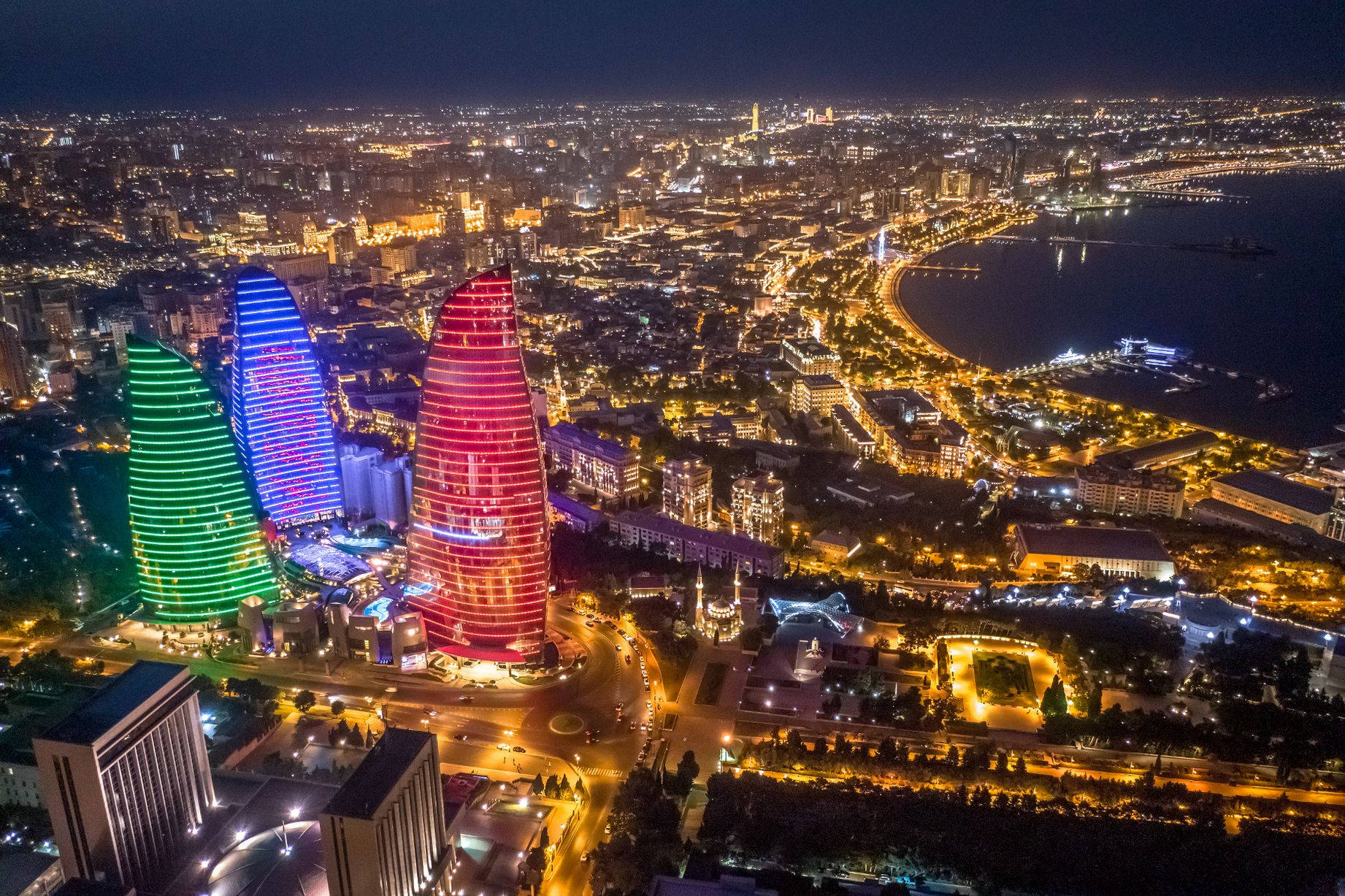 Azerbaijan 桌布 壁紙 — 巴庫火焰塔的夜間航拍視圖，塔身發出紅、藍、綠光，映襯著金色街燈照亮的城市景觀。