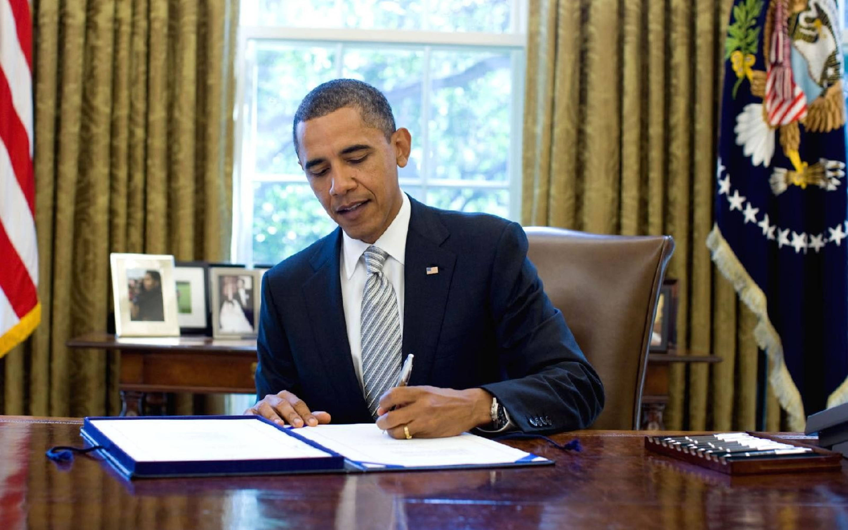 Barack 桌布 壁紙 — 巴拉克·歐巴馬在其總統辦公桌上簽署文件，背景有美國國旗和裱框照片。