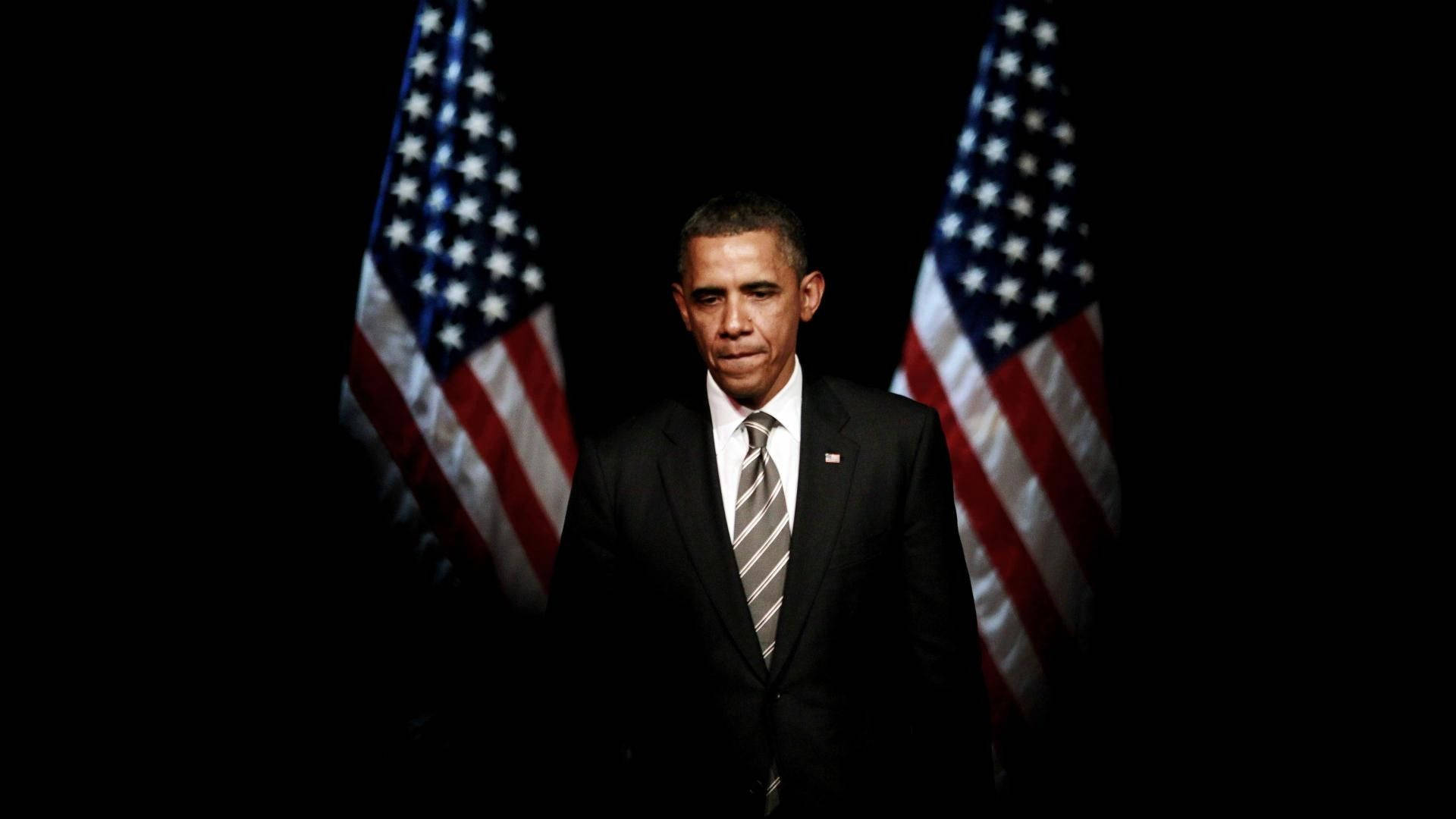 Barack Obama 桌布 壁紙 — 巴拉克·歐巴馬在黑暗的房間中莊嚴地站立，兩側有兩面美國國旗，在戲劇性的聚光燈下。