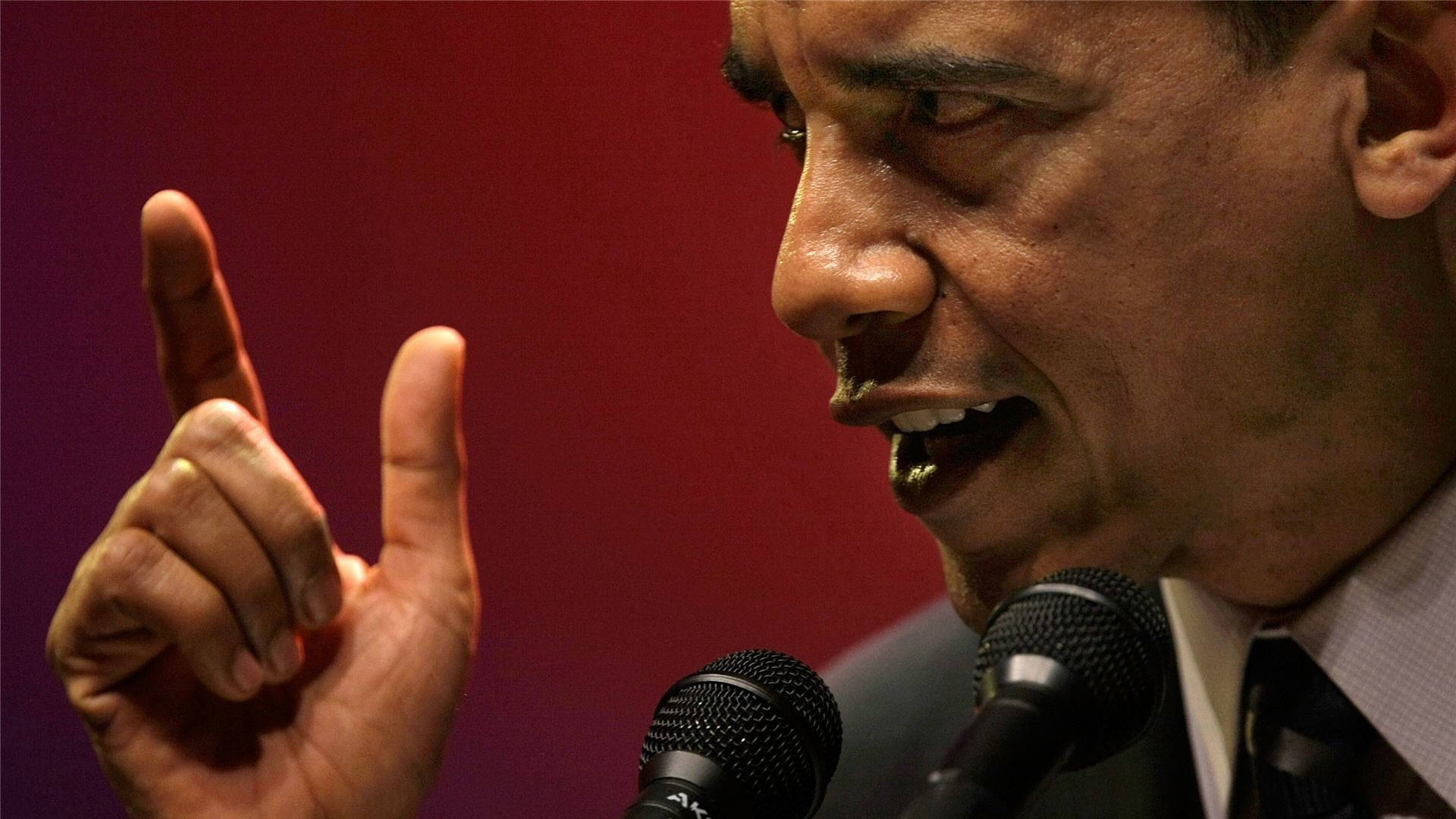 Barack 桌布 壁紙 — 巴拉克·歐巴馬近距離對著麥克風說話，手指豎起，背景為深紅色。