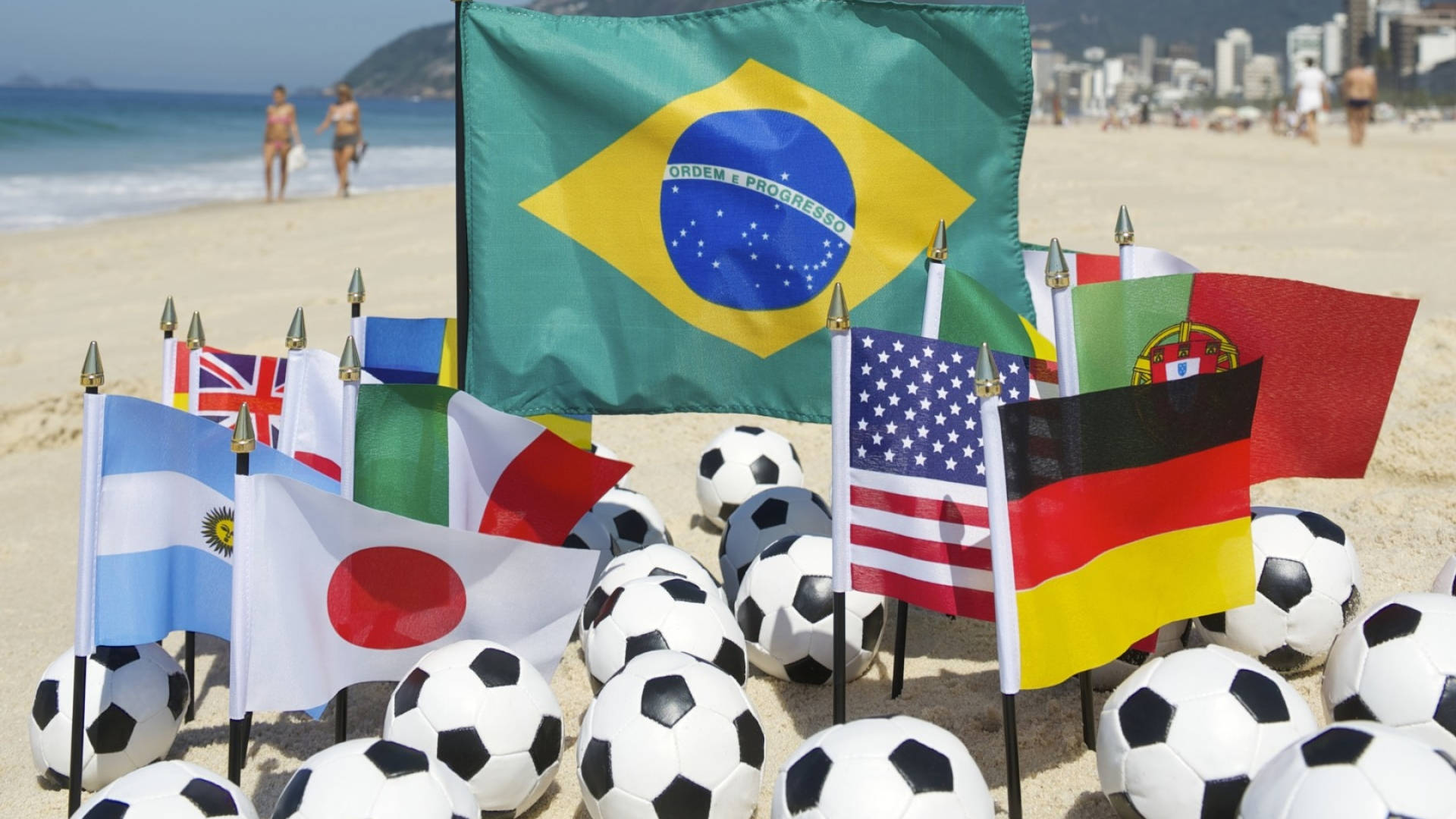 Brazil Flag 桌布 壁紙 — 巴西國旗和國際足球旗幟環繞著黑白足球，位於陽光明媚的海灘上，背景是海洋和城市景觀。