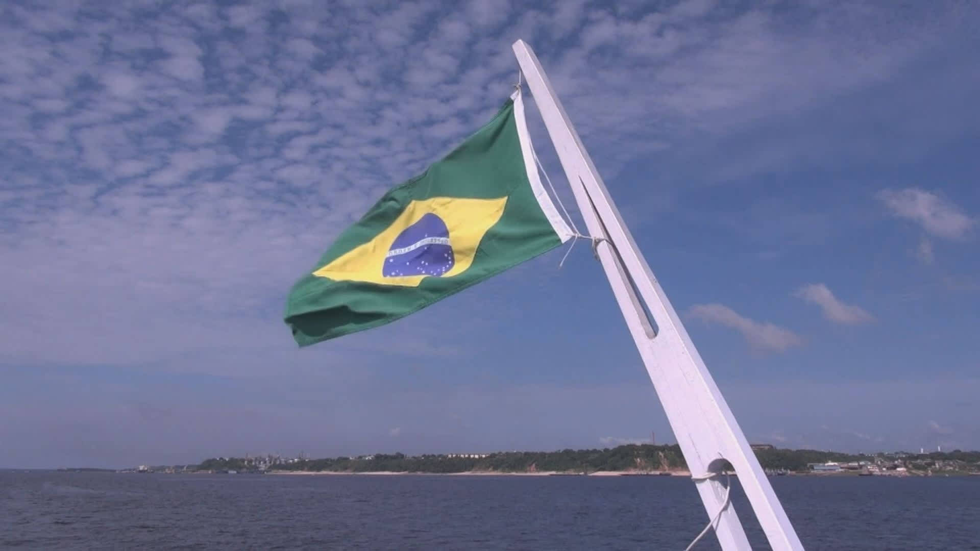 Brazil Flag 桌布 壁紙 — 巴西國旗在船桅上自豪地飄揚，背景是藍天、散落的雲朵和遠處的海岸線。