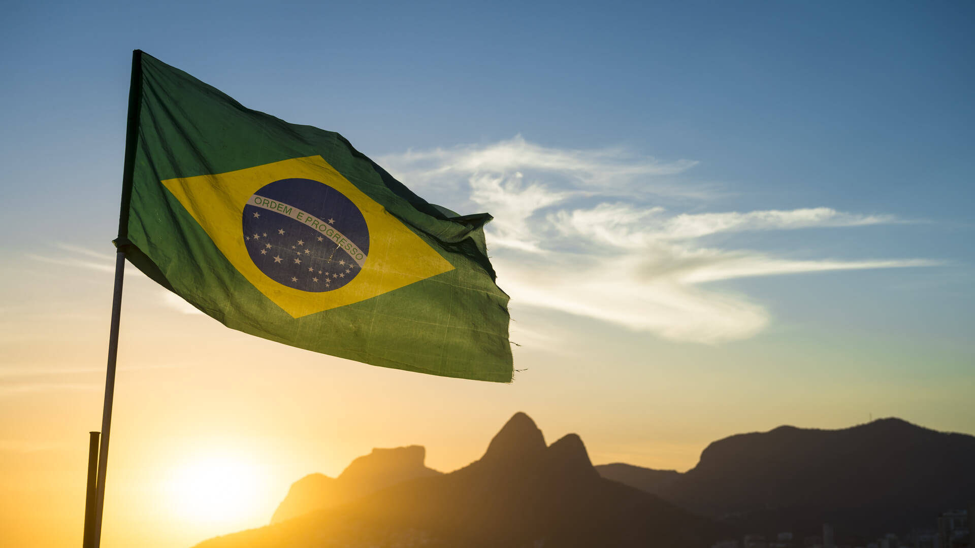 Brazil Flag 桌布 壁紙 — 巴西國旗在金色夕陽天空下自豪地飄揚，背景是輪廓分明的山脈。