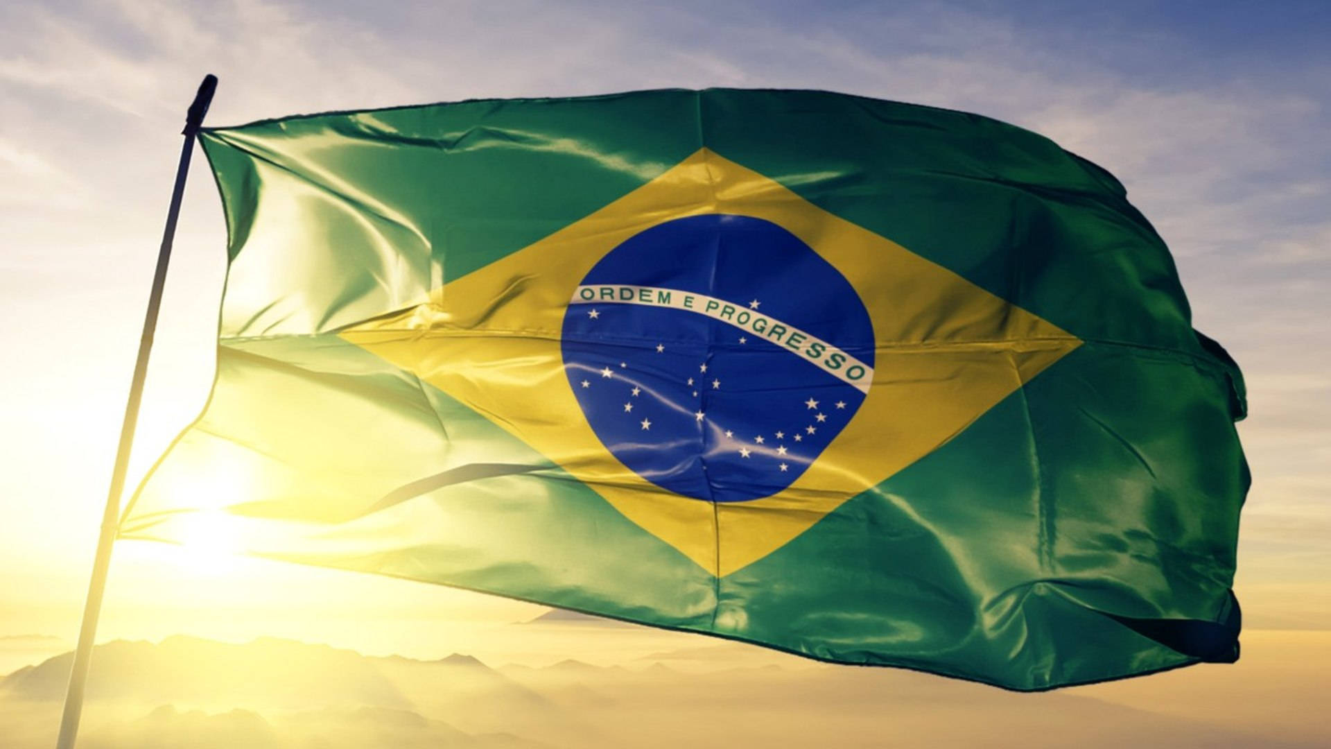 Brazil Flag 桌布 壁紙 — 巴西國旗在金色日出天空下自豪地飄揚，下方有柔軟的雲朵和遠山。