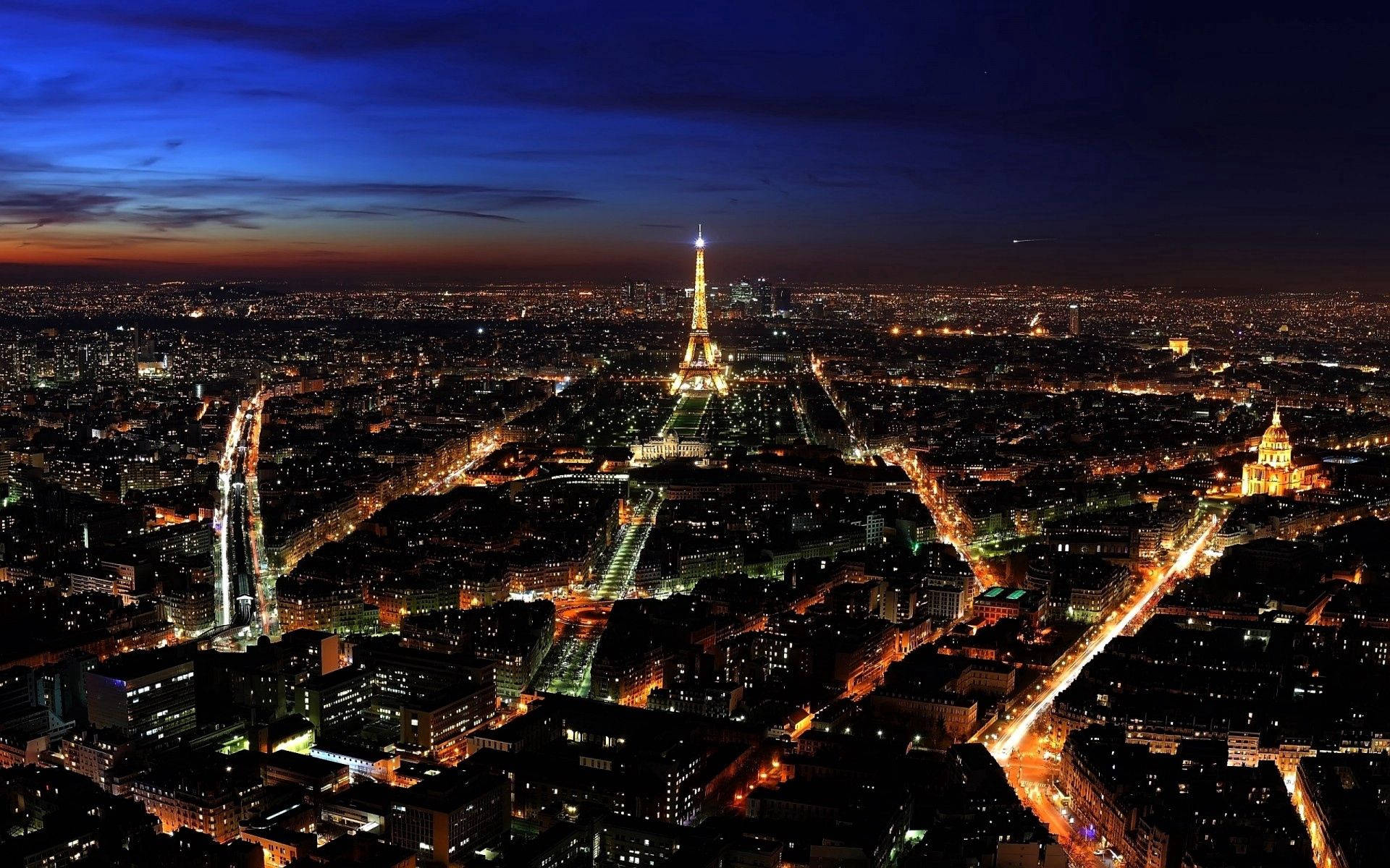 France 桌布 壁紙 — 巴黎天際線在黃昏時分，艾菲爾鐵塔在深藍色天空下照亮了街道和建築物。