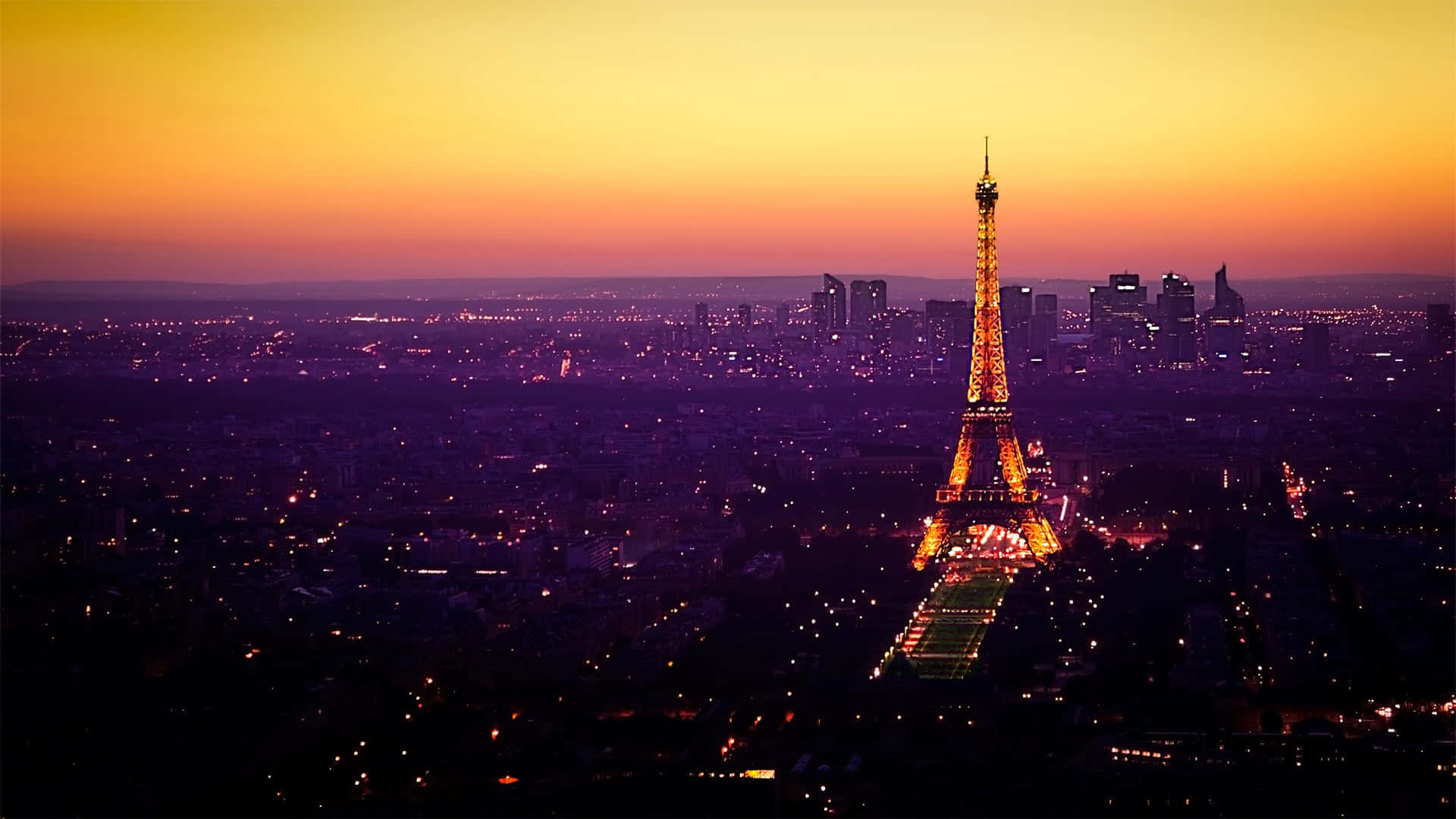Eiffel Tower 桌布 壁紙 — 巴黎日落天際線，艾菲爾鐵塔在橙色和紫色漸變的天空中閃爍琥珀色光芒。
