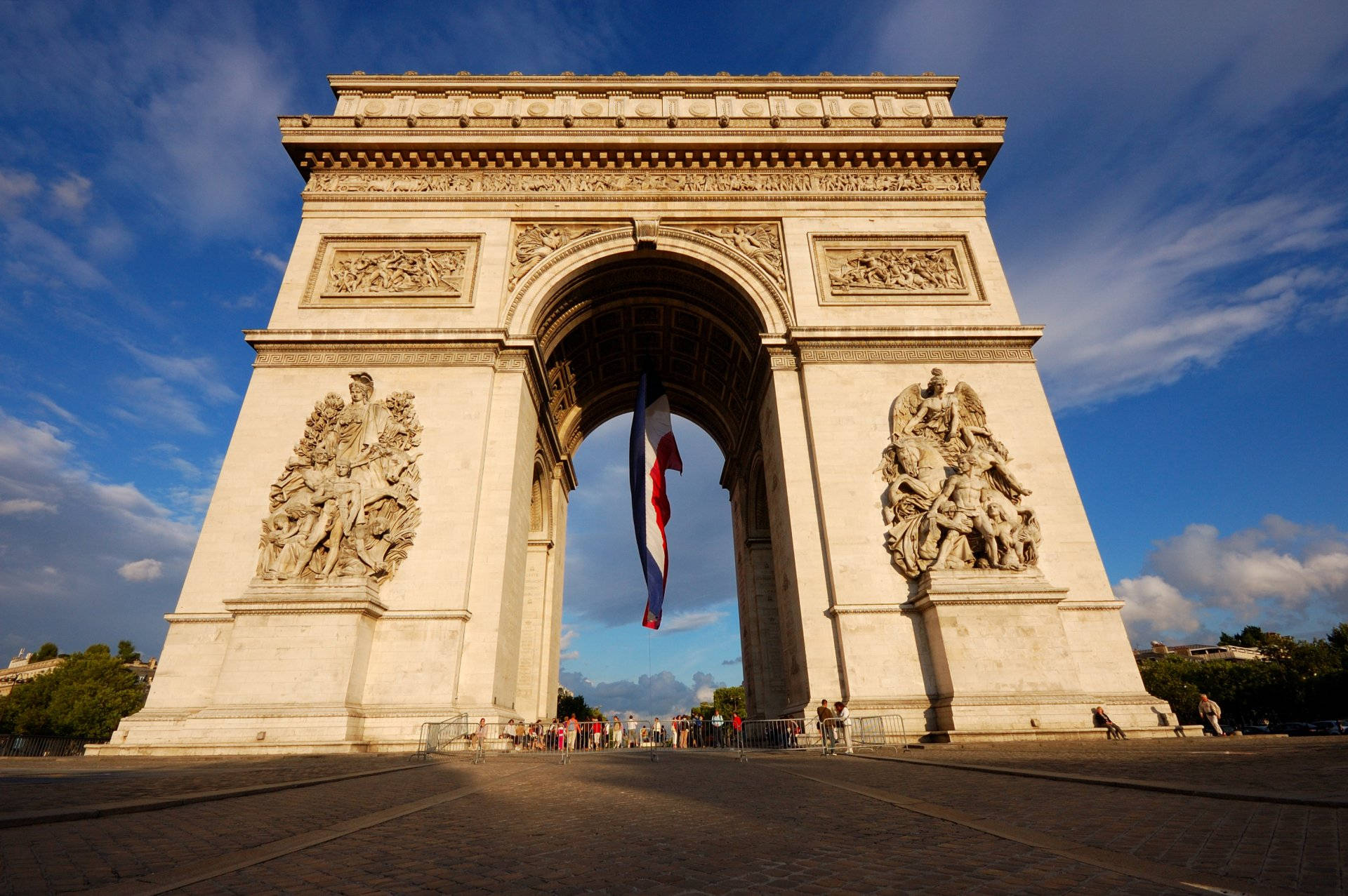 France Flag 桌布 壁紙 — 巴黎的凱旋門在湛藍的天空、散落的雲朵和懸掛在拱門上的法國國旗下高高聳立。