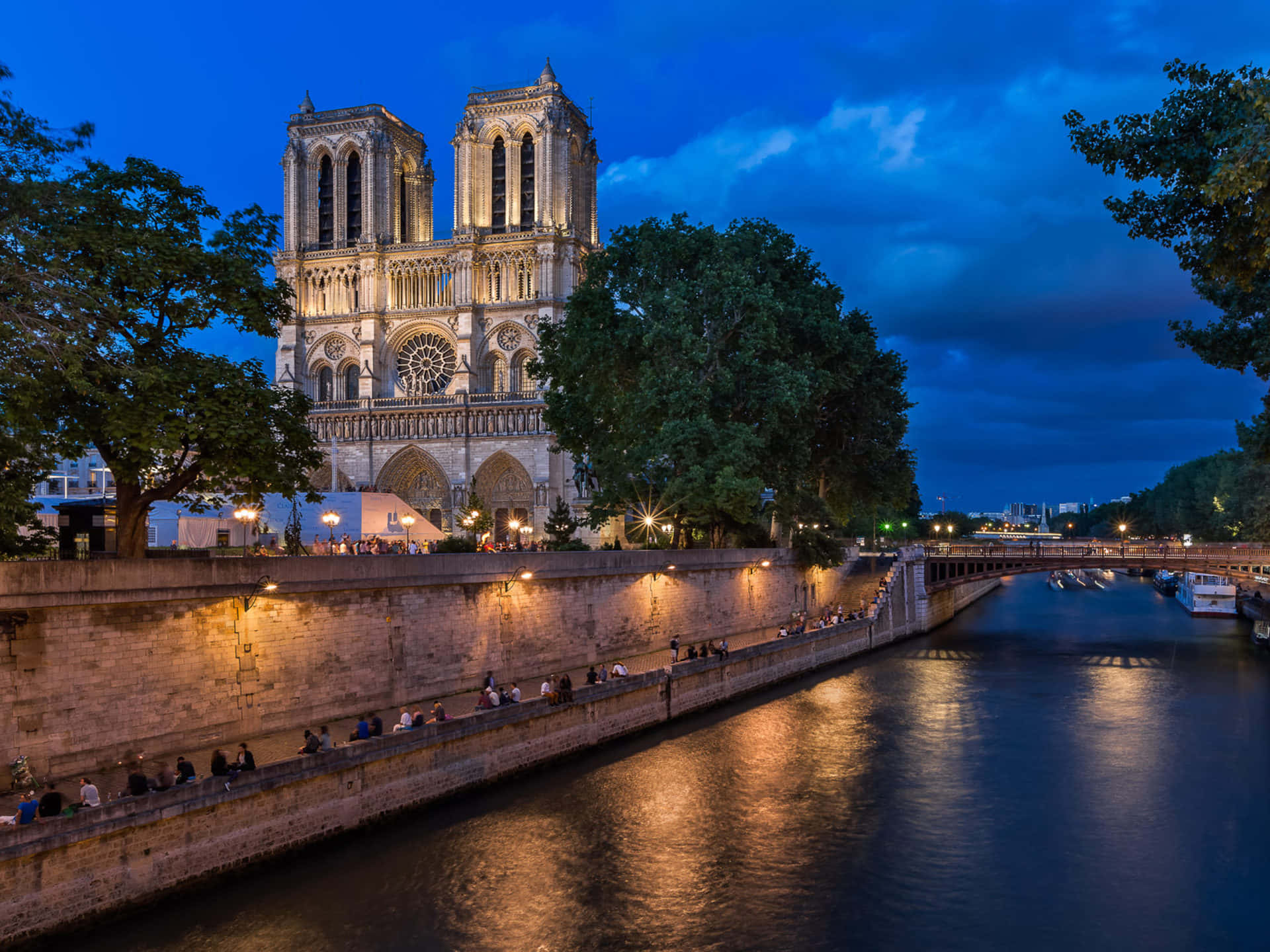 Notre Dame 桌布 壁紙 — 巴黎聖母院在深藍色的暮色天空中溫暖地發光，人們沿著塞納河畔放鬆。
