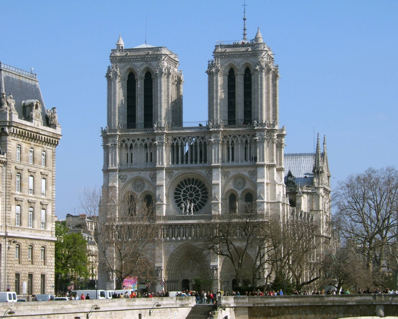 Notre Dame 桌布 壁紙 — 巴黎聖母院在清澈的藍天下雄偉矗立，周圍有光禿的樹木和聚集在底部的遊客。