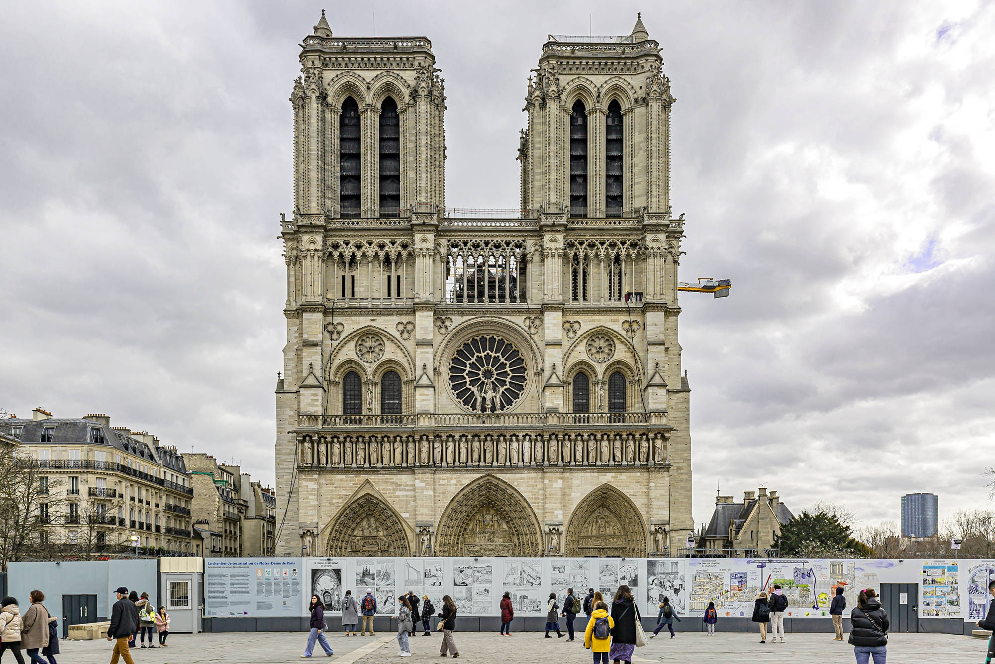 Notre Dame 桌布 壁紙 — 巴黎聖母院在陰雲密佈的天空下巍然聳立，遊客在其標誌性哥德式立面前行走。