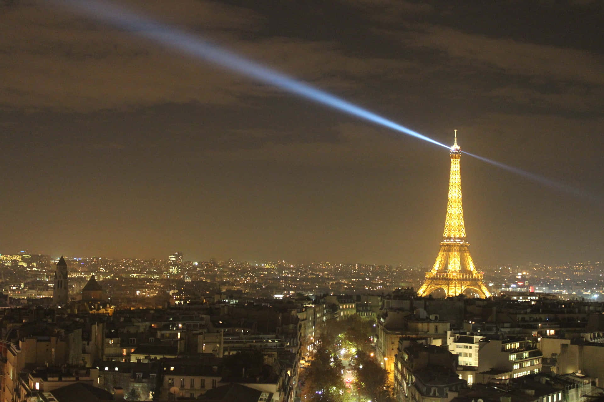 Eiffel Tower 桌布 壁紙 — 巴黎鐵塔在夜晚閃耀著金色光芒，一道明亮的藍色雷射光束劃過巴黎上空多雲的天空。