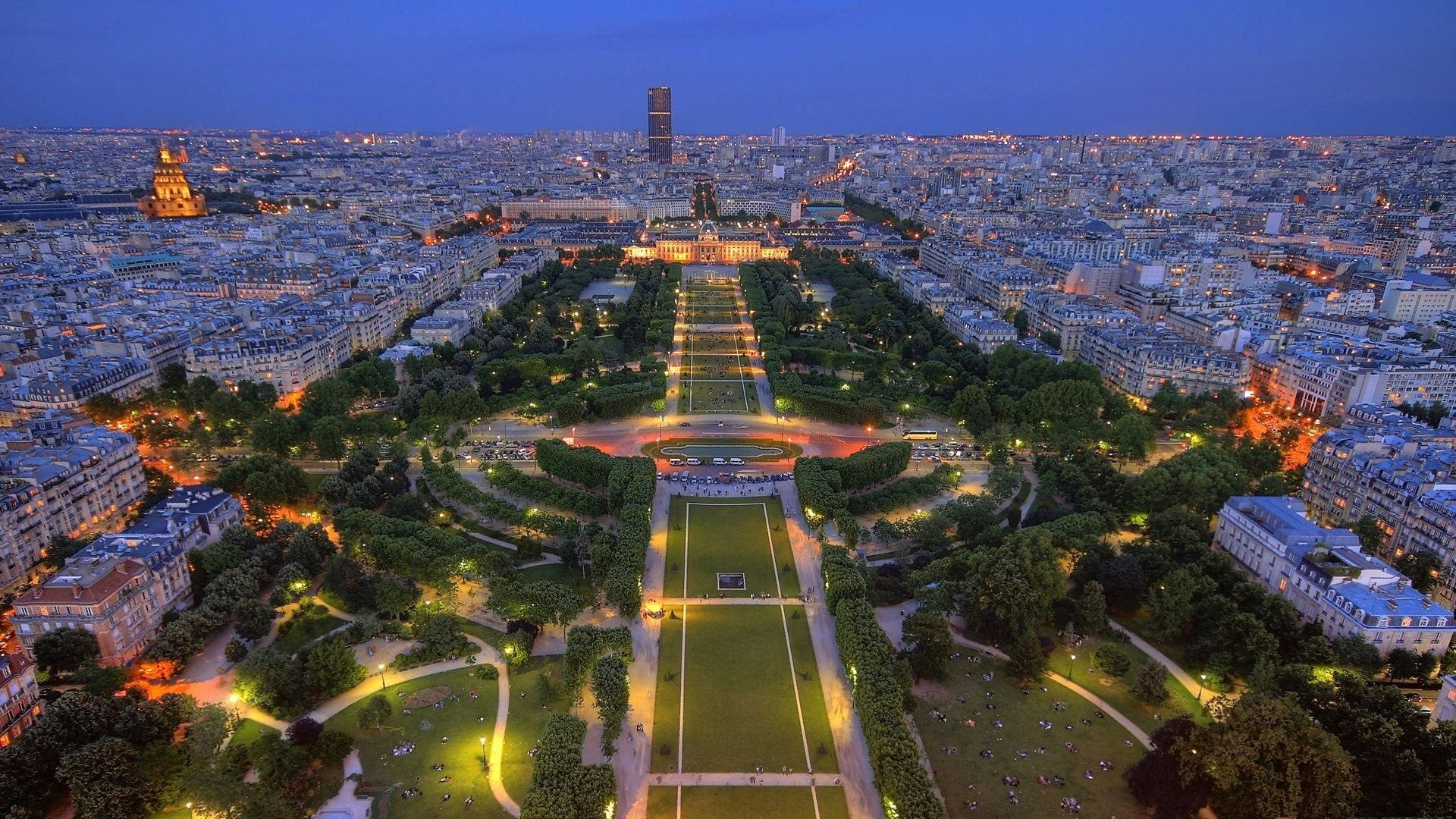 France 桌布 壁紙 — 巴黎黃昏時的鳥瞰圖，展示了燈火通明的杜樂麗花園和周圍的城市景觀，伴隨著閃爍的燈光和漸暗的天空。