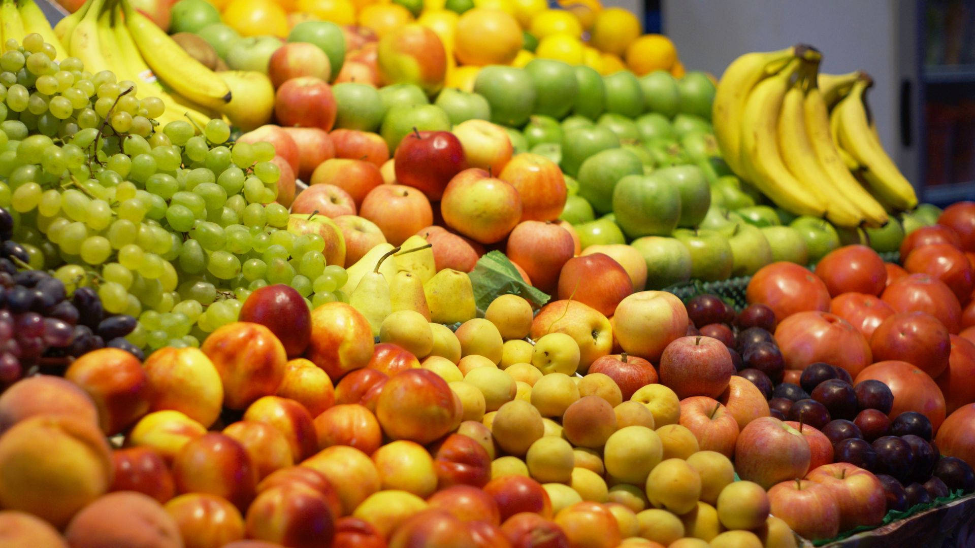 Fruits 桌布 壁紙 — 市場陳列中各種新鮮水果（如香蕉、葡萄、蘋果和李子）高高堆起的鮮豔特寫。