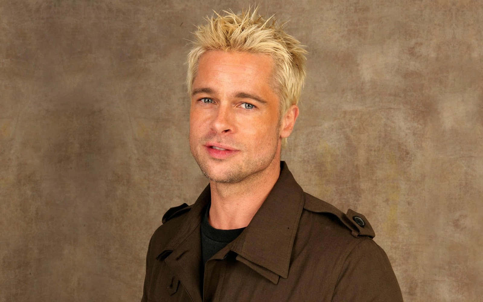 布萊德彼特 桌布 壁紙 — 布萊德彼特（Brad Pitt）留著尖刺金髮和藍眼睛，身穿棕色風衣，背景為有紋理的米色背景。