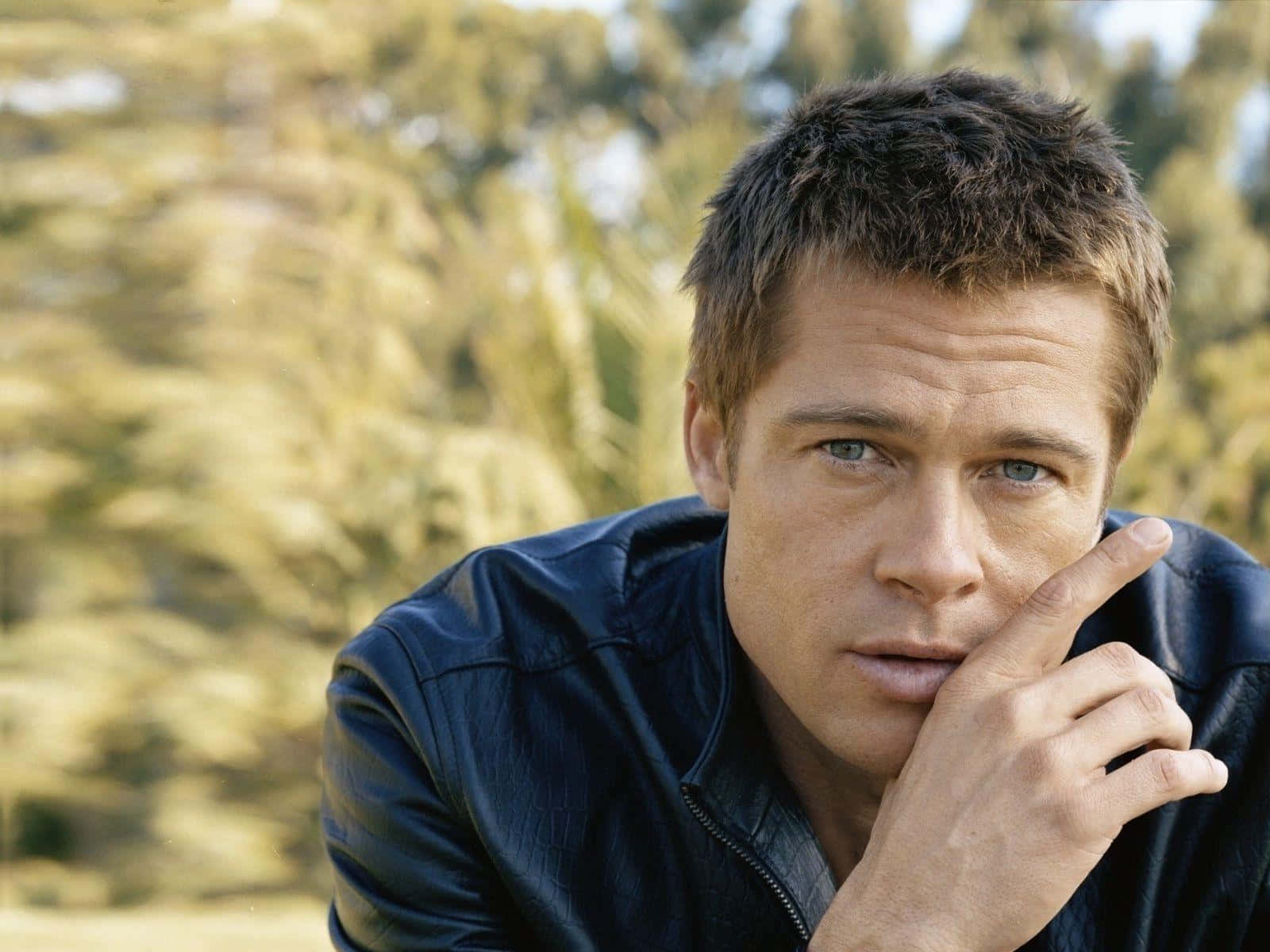 Brad Pitt 桌布 壁紙 — 布萊德彼特穿著黑色皮夾克，手托下巴，背景是陽光照射下模糊的樹葉。