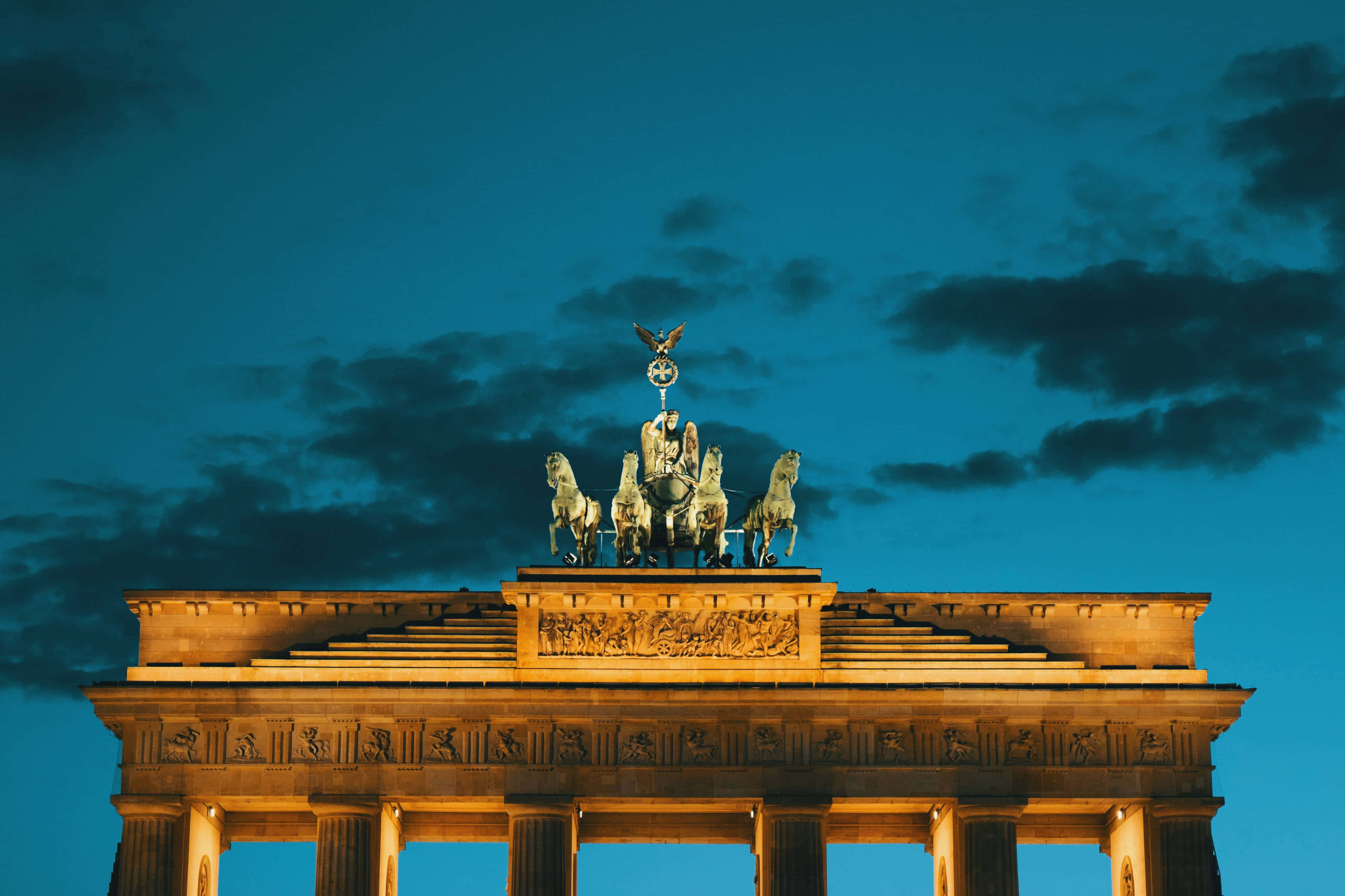 Berlin 桌布 壁紙 — 布蘭登堡門在黃昏時亮起，其四馬戰車雕像在深藍色天空和散落的雲朵中閃耀。
