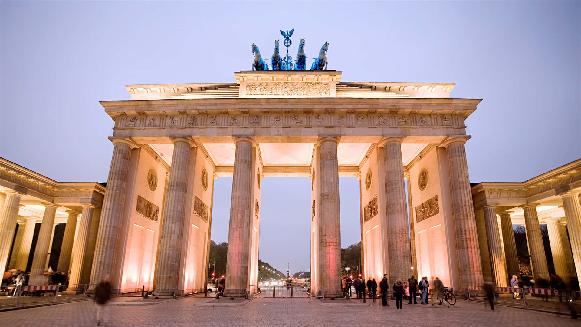 Berlin 桌布 壁紙 — 布蘭登堡門在黃昏時亮起，溫暖的燈光在其新古典主義柱子和上方的四馬戰車雕像上投下金色光輝。