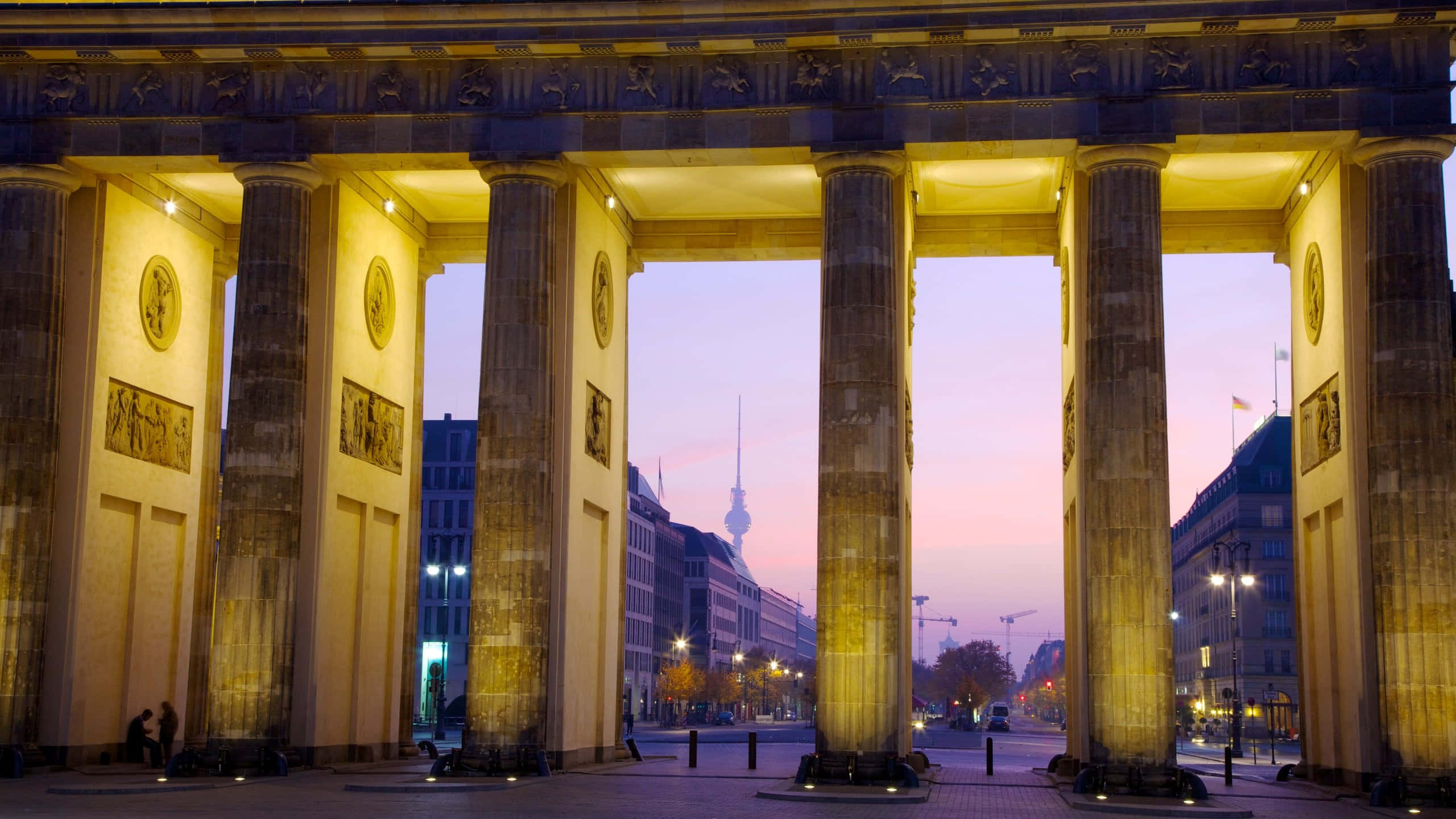 Berlin 桌布 壁紙 — 布蘭登堡門在黃昏時亮起，溫暖的燈光在其新古典主義柱子上投射出金色光芒，框住了遠處有電視塔的城市景觀。