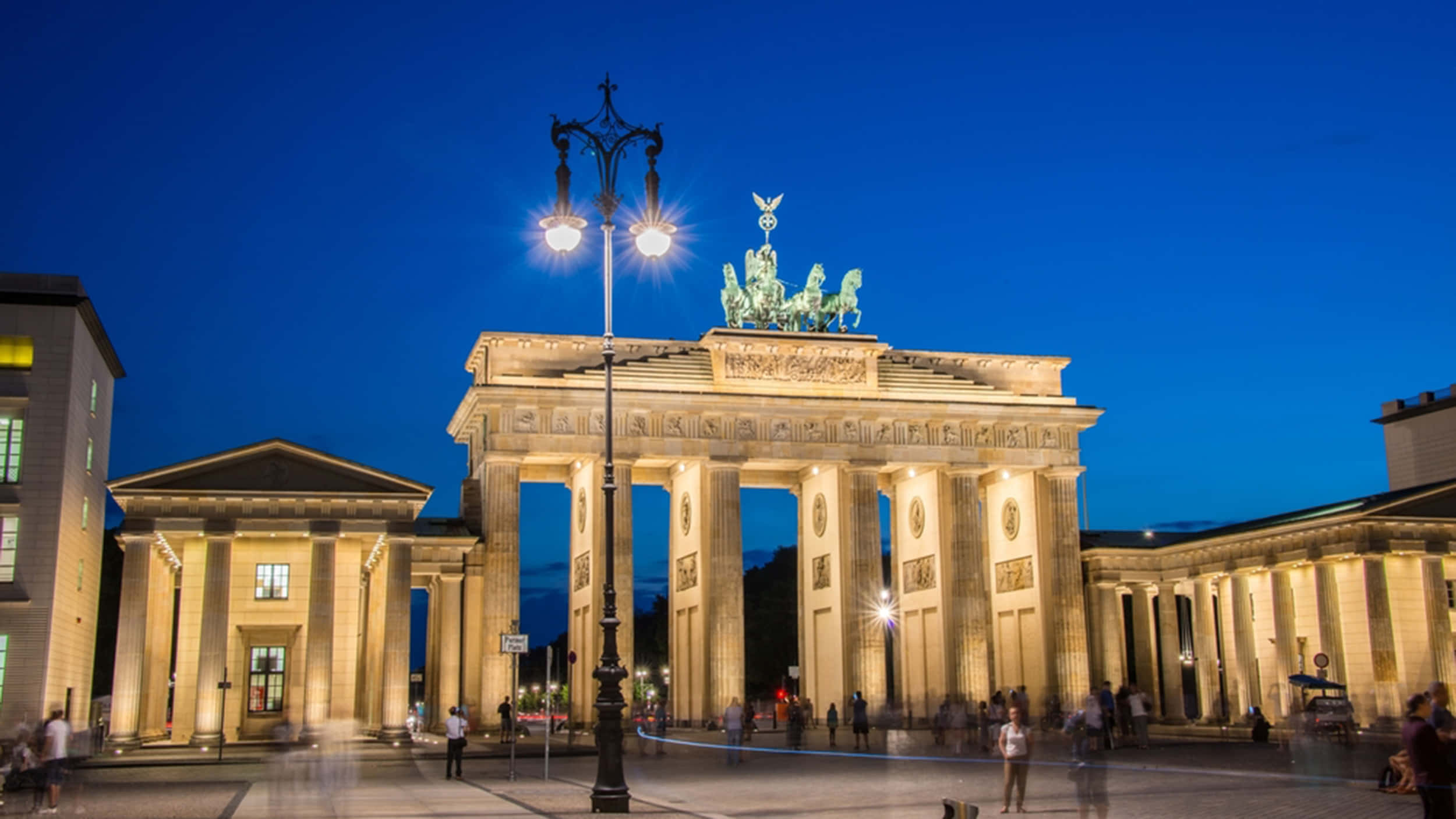 Berlin 桌布 壁紙 — 布蘭登堡門在黃昏時被溫暖的燈光照亮，襯托著深藍色的天空和下方模糊的行人。