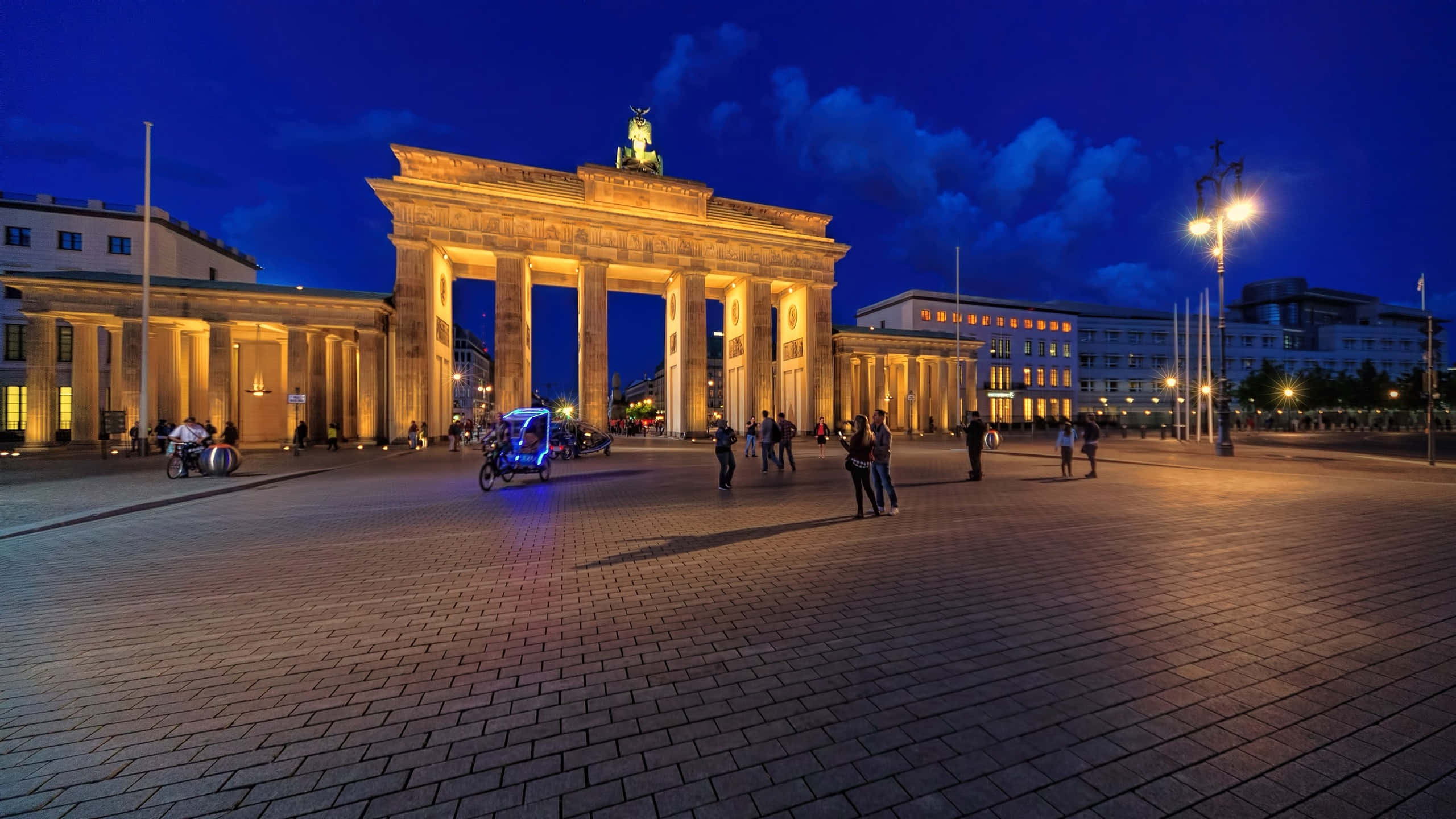 Berlin 桌布 壁紙 — 布蘭登堡門在黃昏時被溫暖的燈光照亮，在鵝卵石廣場上投下長長的影子，人們漫步其中，一輛發光的三輪車駛過。
