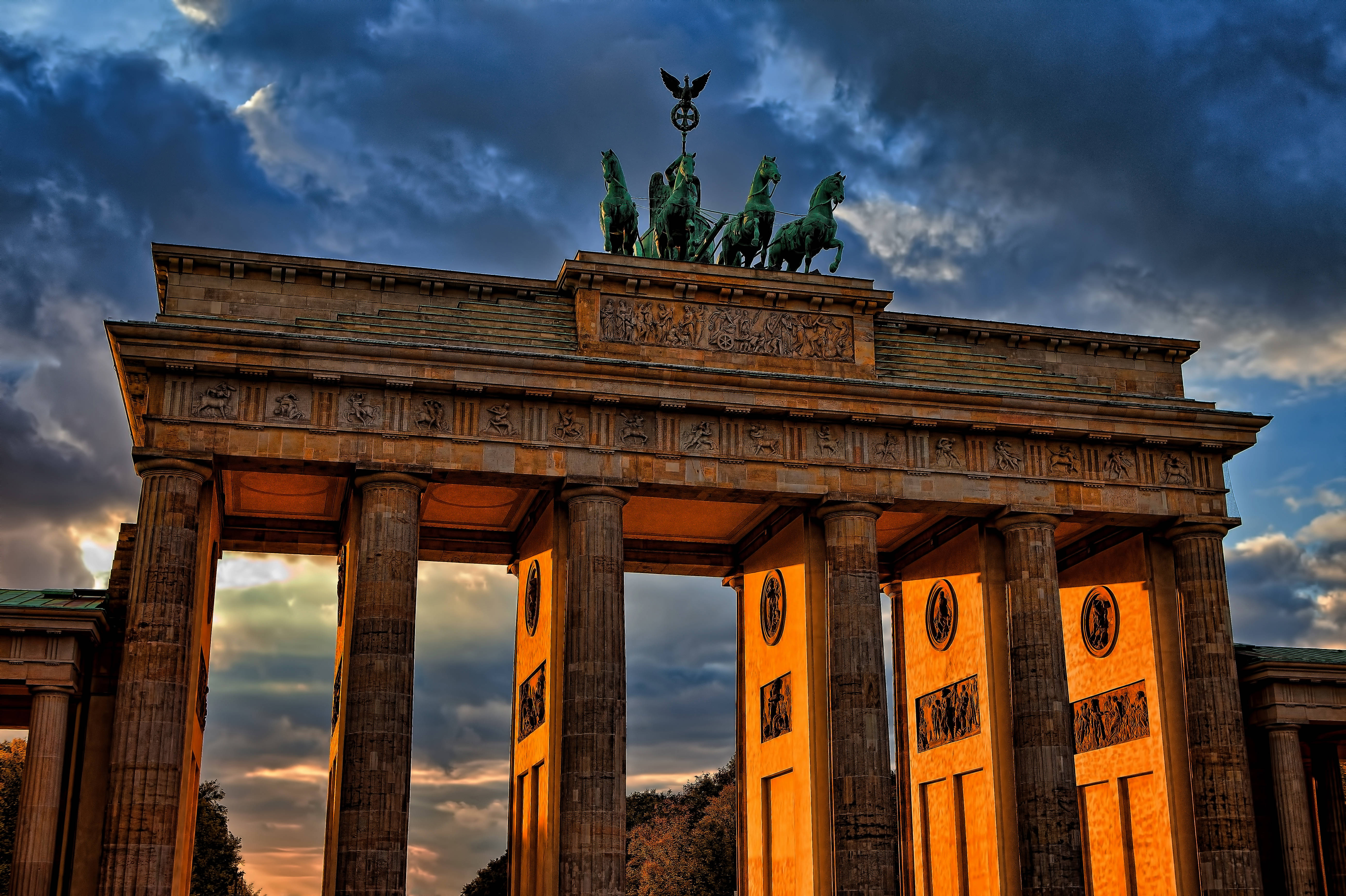 Berlin 桌布 壁紙 — 布蘭登堡門沐浴在金色的夕陽光芒中，其新古典主義柱子和四馬戰車雕像上方有戲劇性的雲彩。
