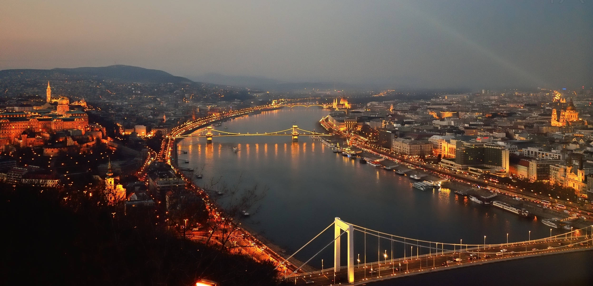 Hungary 桌布 壁紙 — 布達佩斯的全景日落景色，多瑙河上燈火通明的橋樑和歷史建築溫暖地閃耀。